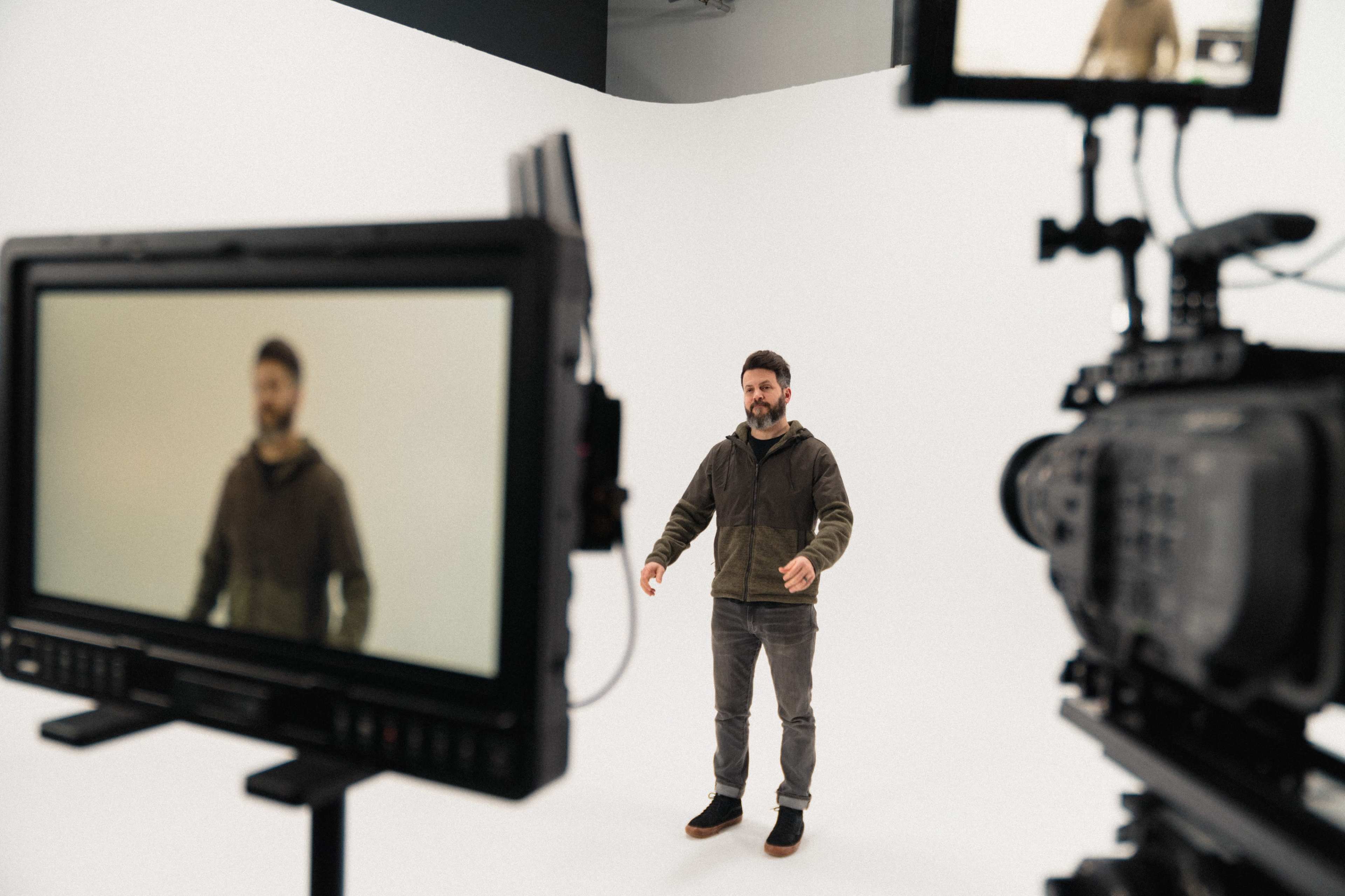 A man stands in front of a white backdrop while being filmed by two cameras.