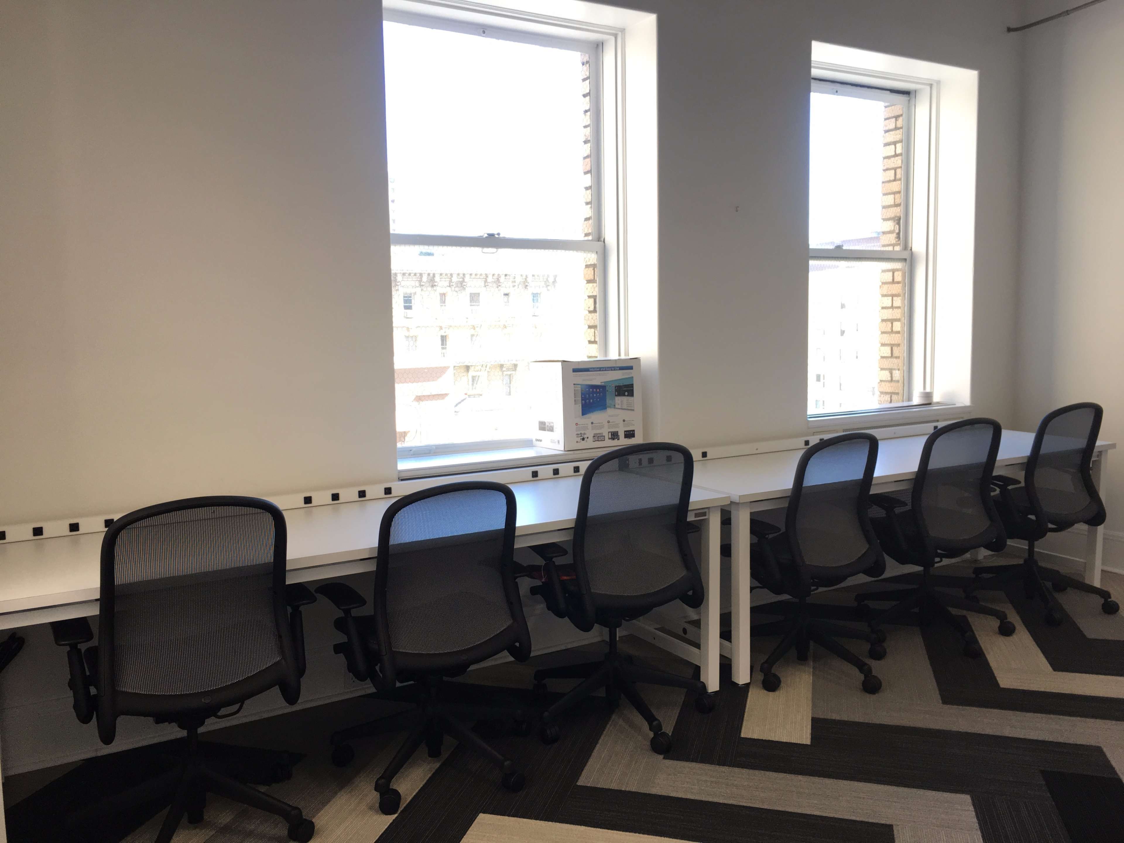 A row of empty office desks with ergonomic chairs is positioned beside large windows in a well-lit workspace.