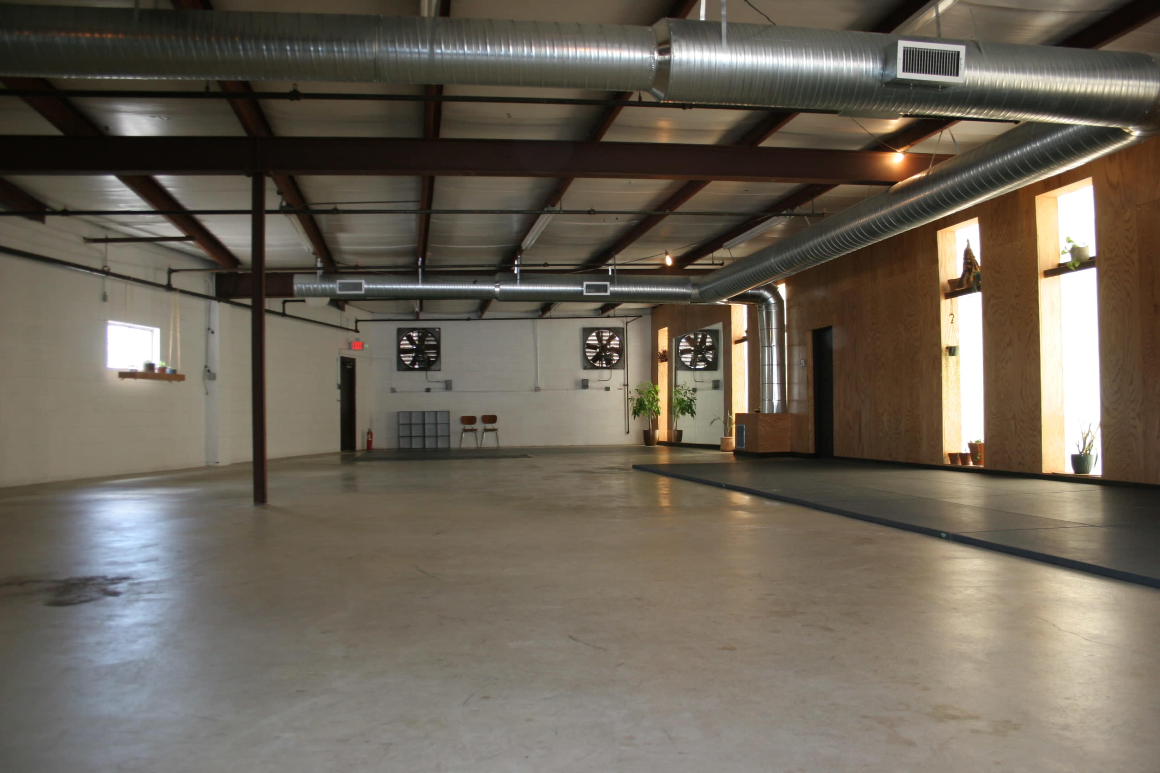 An empty industrial space with concrete flooring, metal beams, and large windows featuring plants.