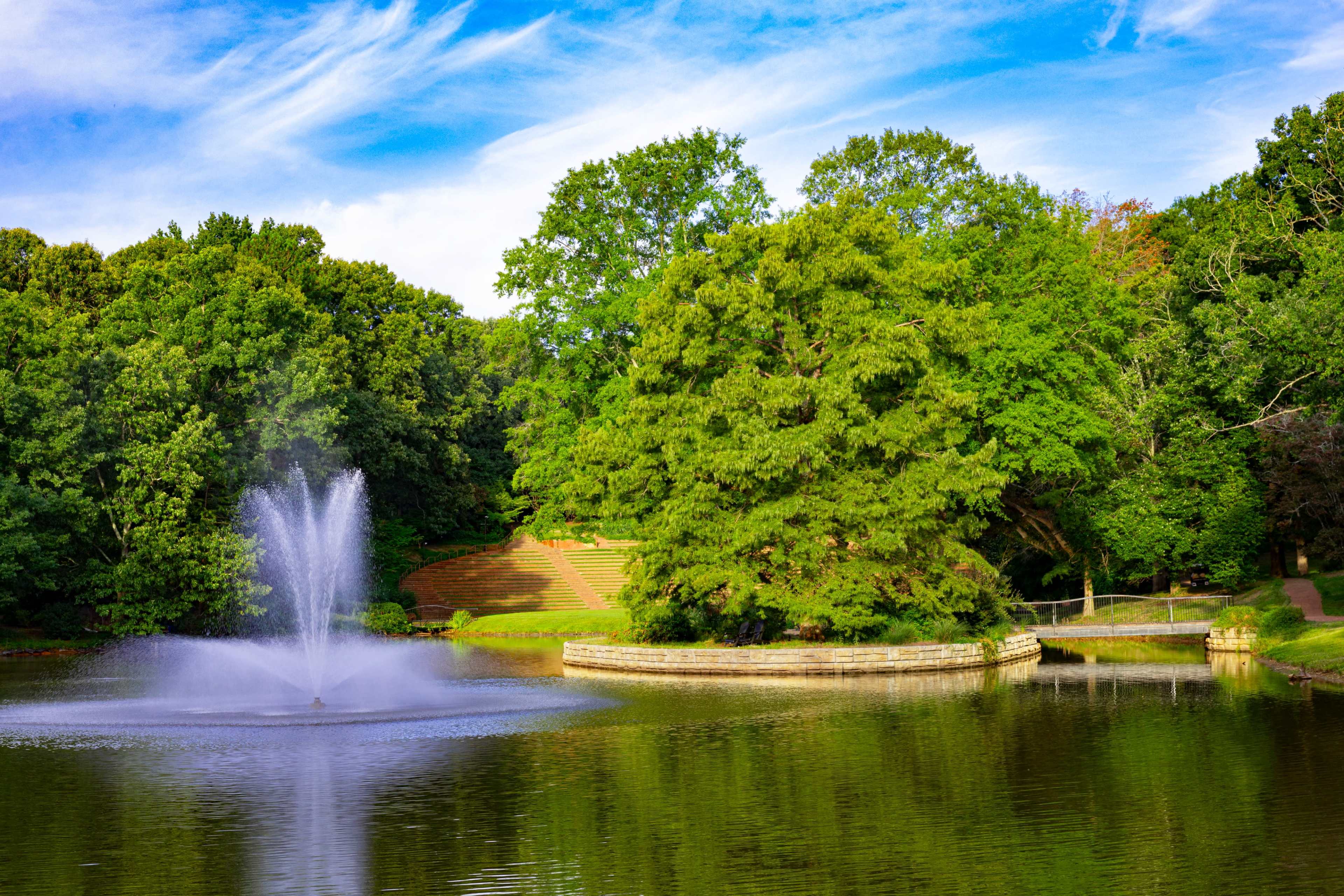 A fountain sprays water in a calm pond surrounded by lush green trees and a landscaped area.