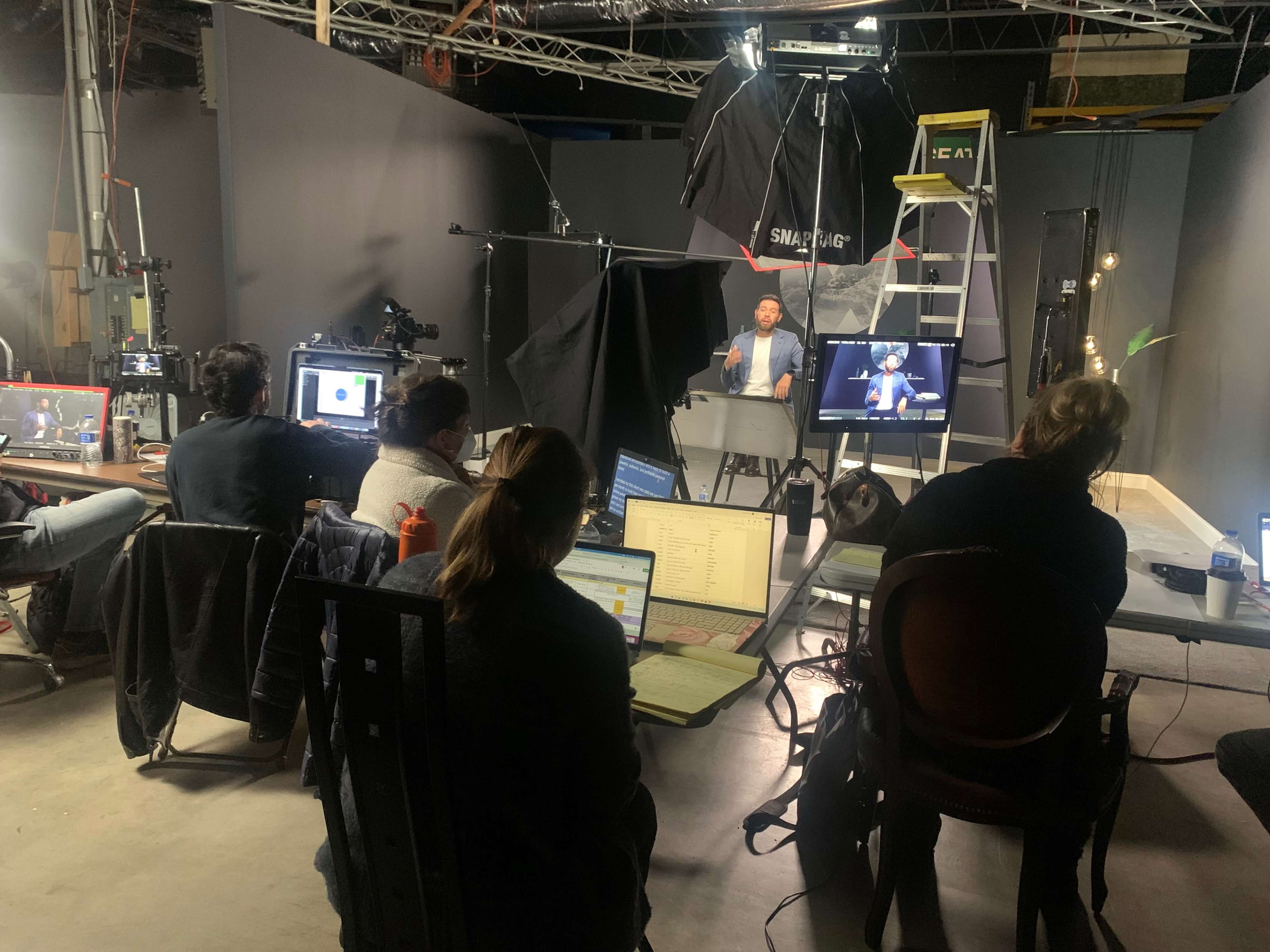 A group of people is seated at tables in a studio, watching a presentation on a screen while surrounded by cameras and lighting equipment.