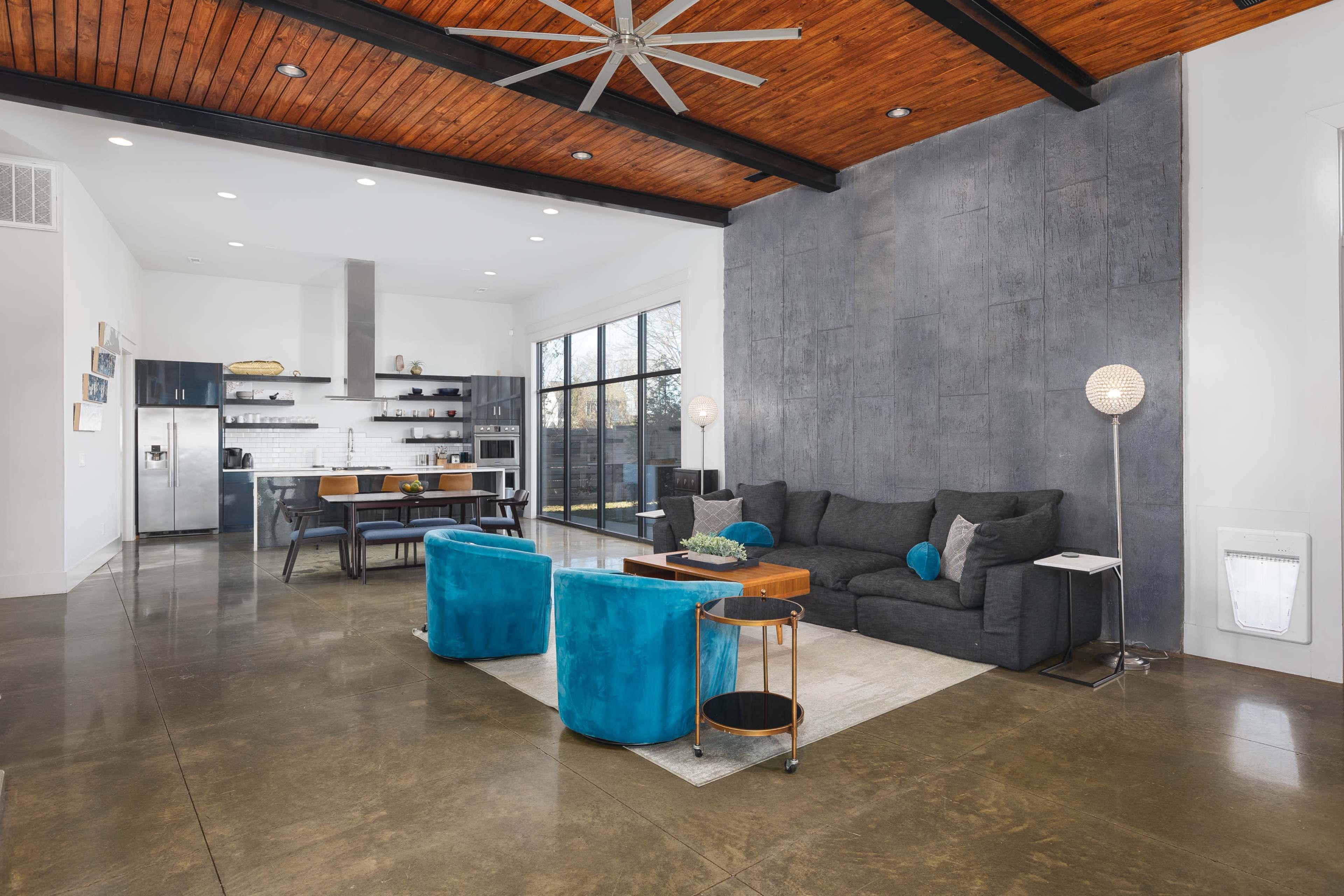 A modern living room featuring a large dark sectional sofa, two vibrant blue chairs, a wooden table, and a kitchen area with sleek appliances.