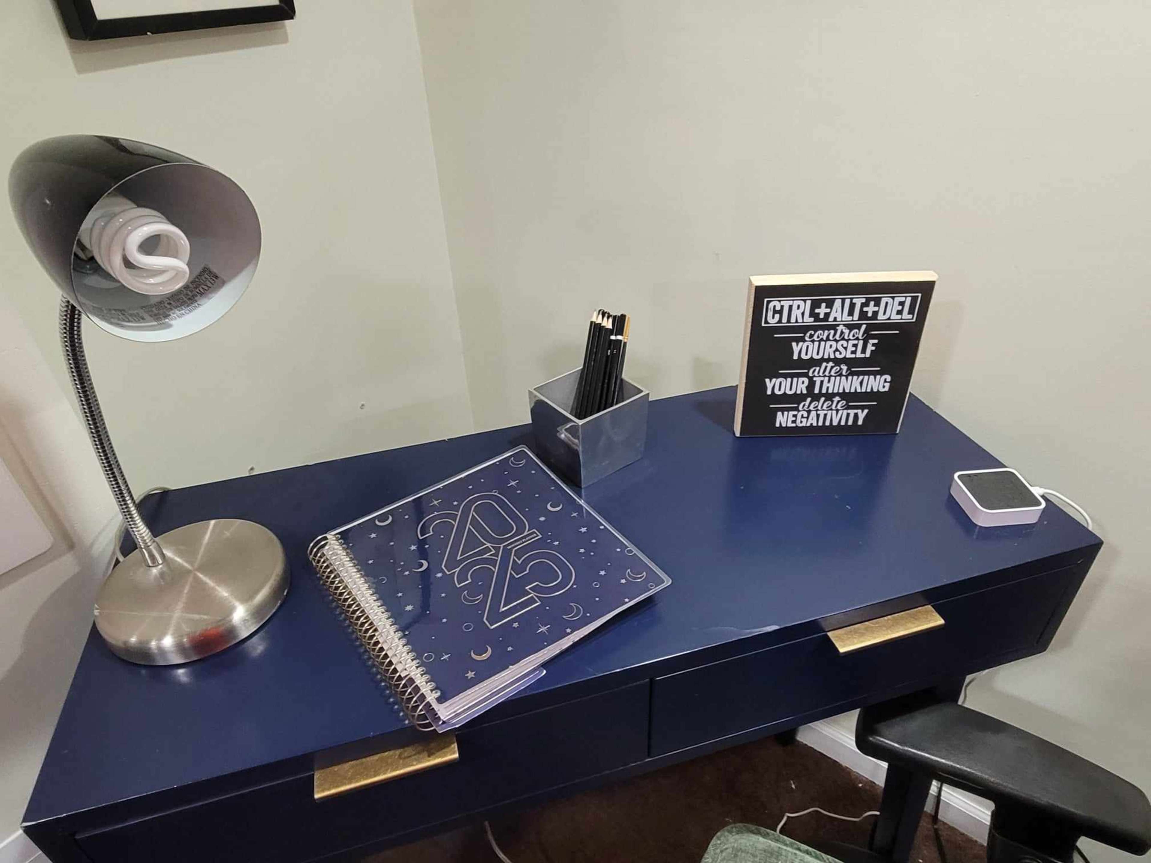 The image shows a desk with a lamp, a container of pens, a planner marked "2025," and a sign with motivational text.
