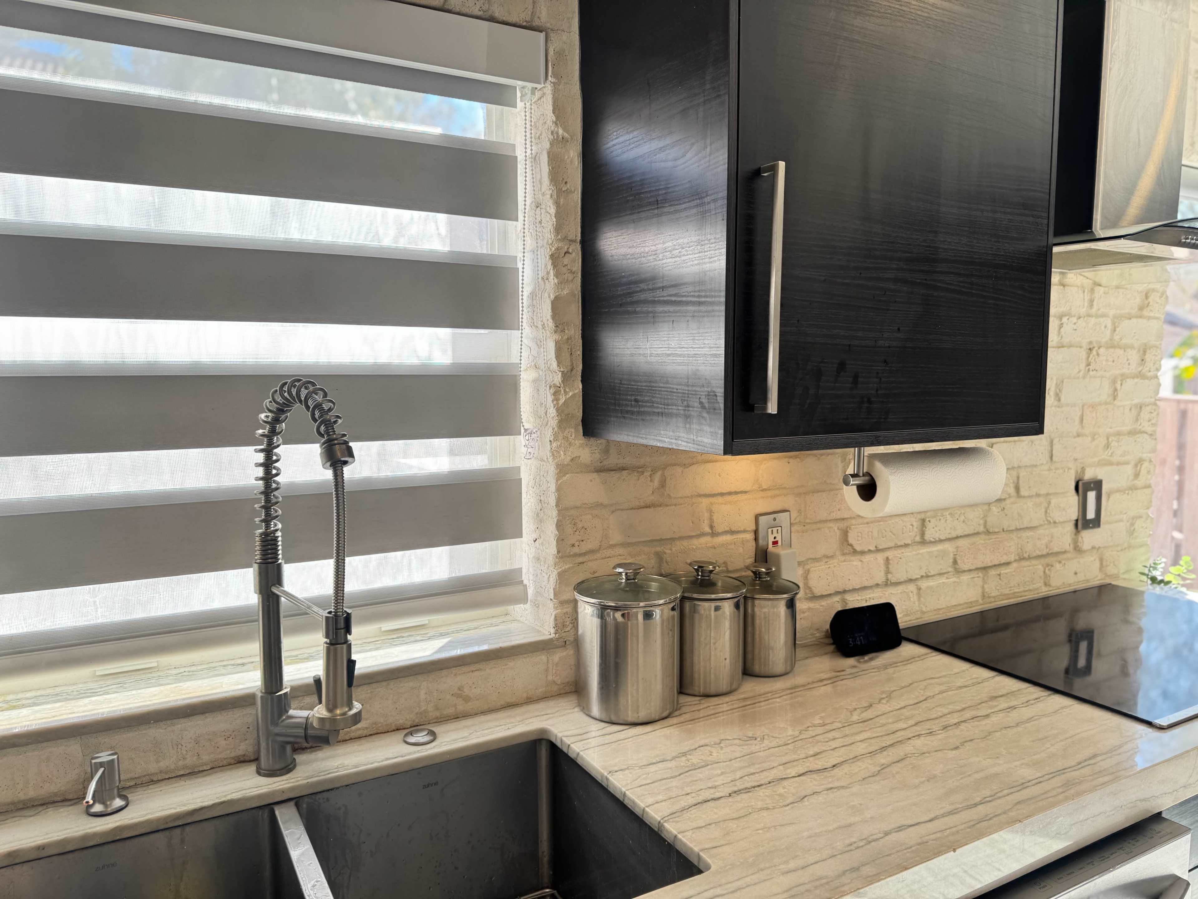 The image shows a modern kitchen sink area with a pull-down faucet, a small digital clock, and several metal containers on a light-colored countertop.