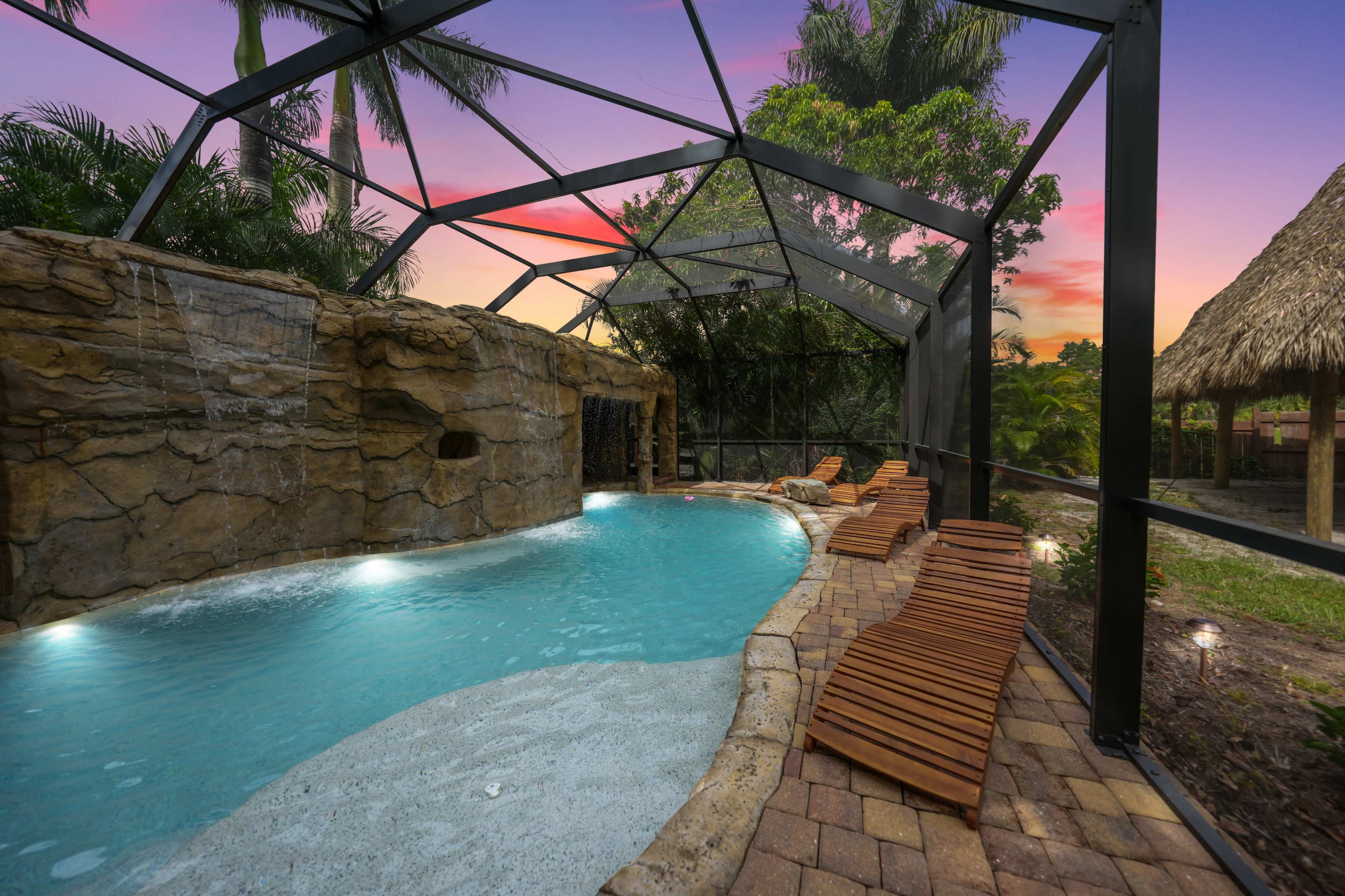 A pool area enclosed by a screened structure, featuring a rock waterfall, wooden lounge chairs, and lush greenery at sunset.