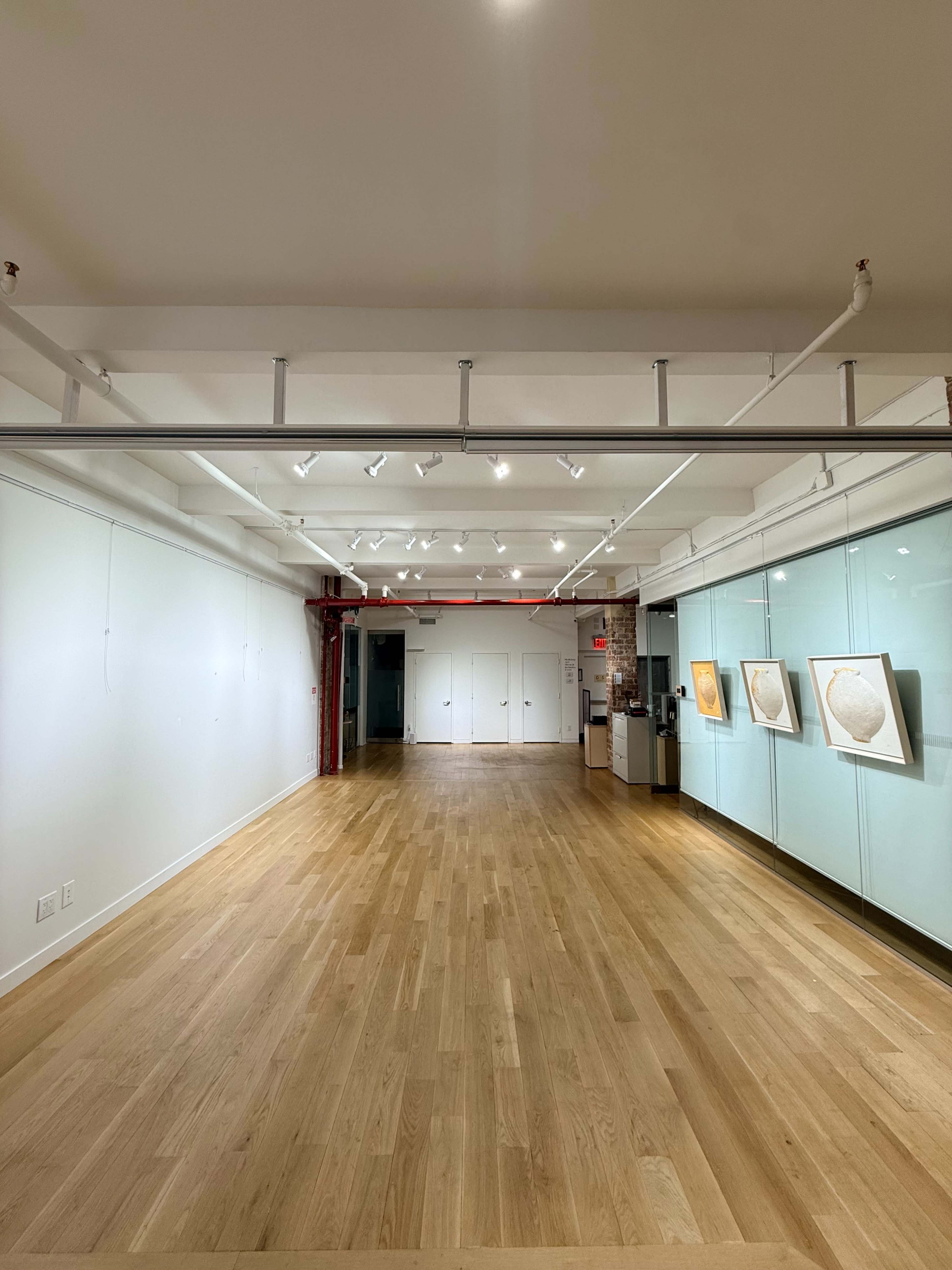 The image shows an empty art gallery with hardwood floors, white walls, and a few framed artworks displayed on one side.