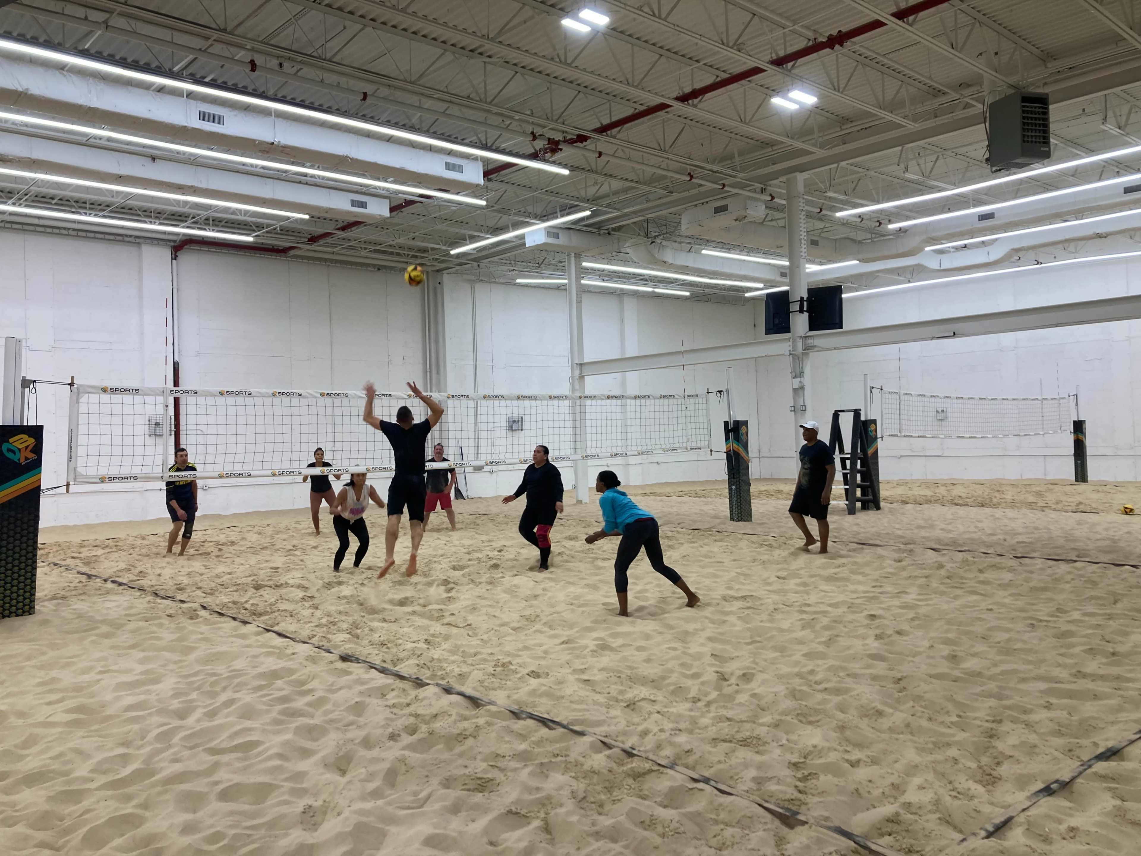 A group of six people play beach volleyball indoors on a sand court.