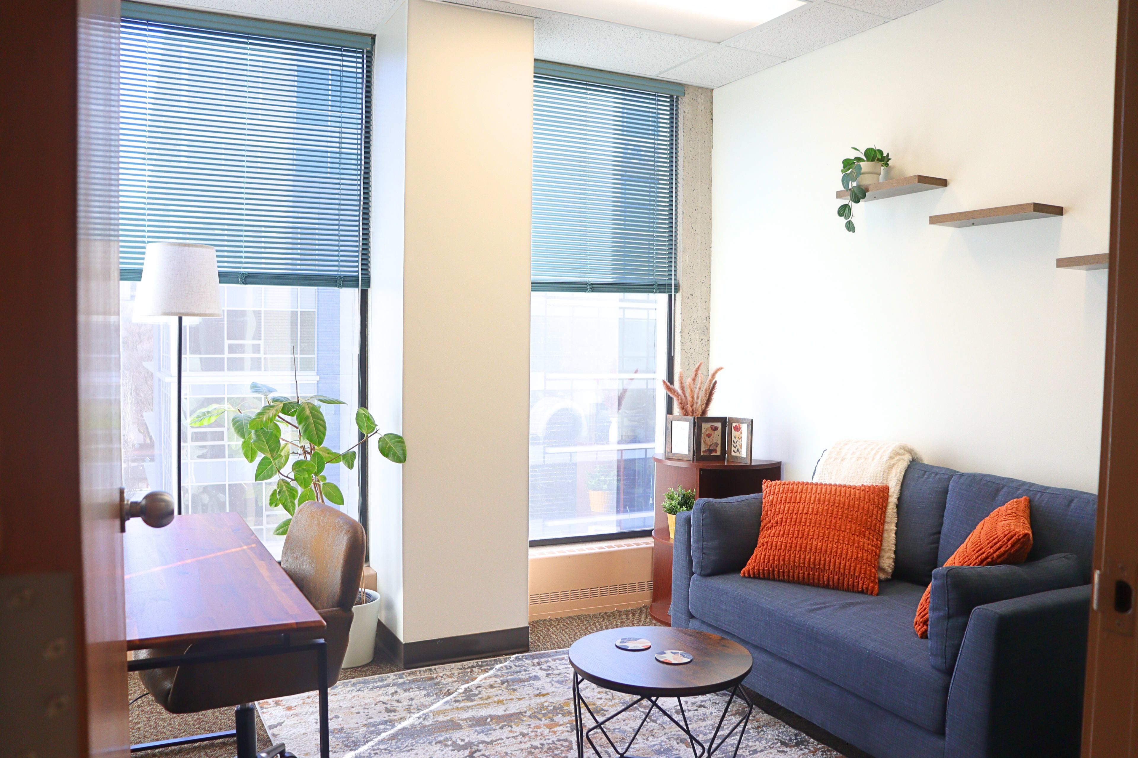 The image displays a small office space with a blue couch, a wooden desk, and two large windows featuring blinds, alongside decorative plants and wall shelves.