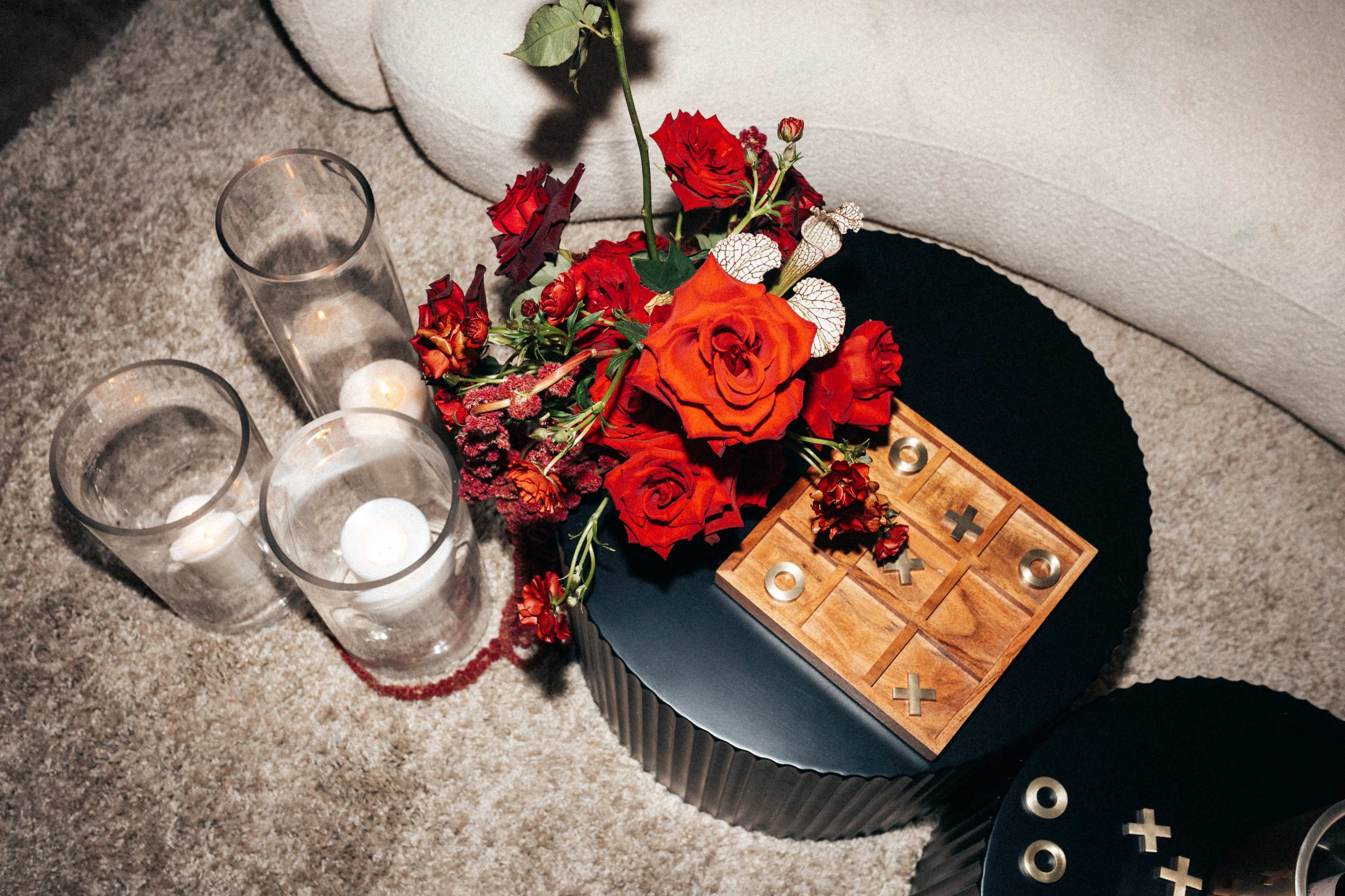 A small round black table holds a wooden tic-tac-toe set and a bouquet of red roses alongside three glass candles.