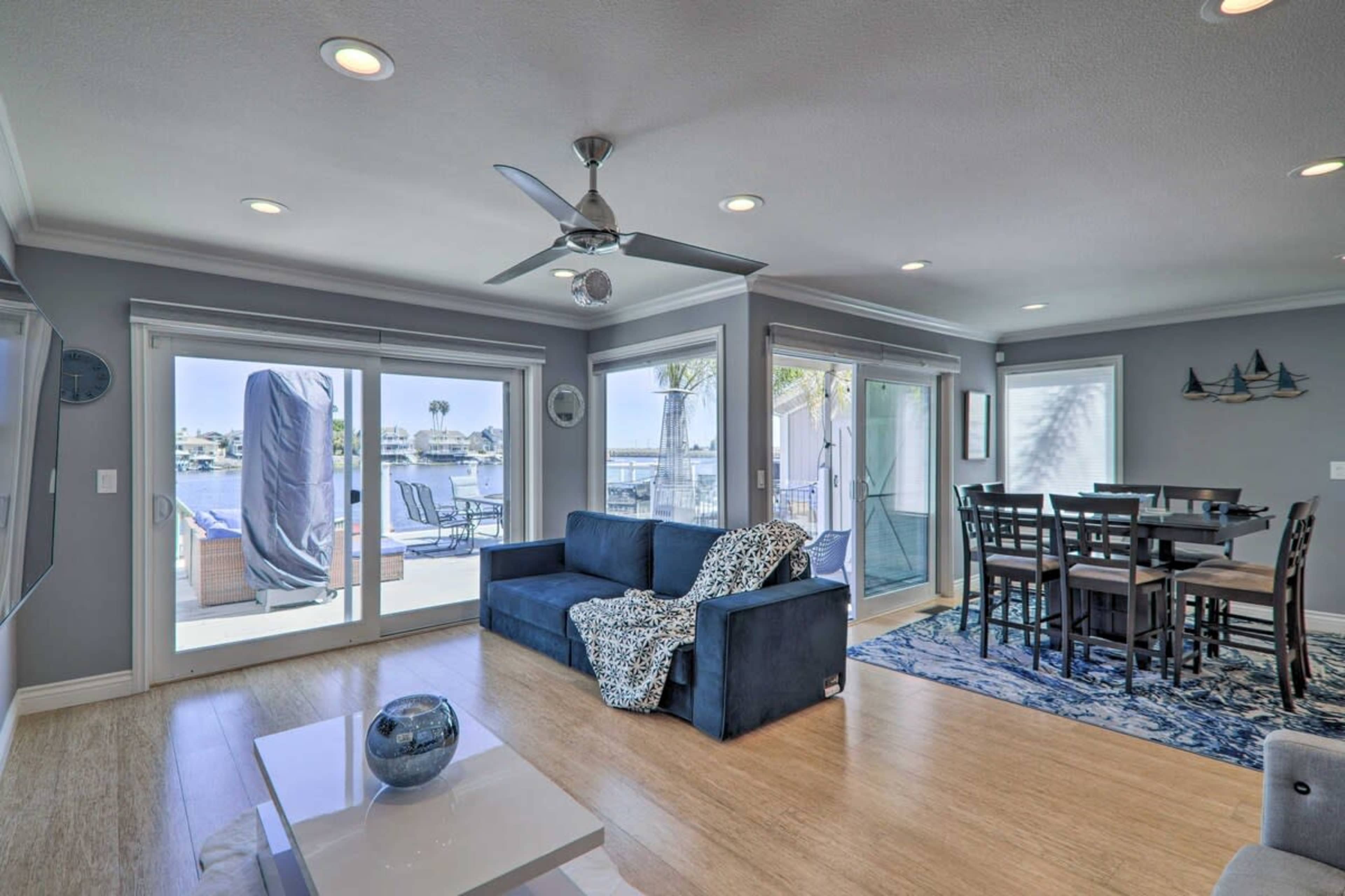 A living room with a sofa, dining area, and large windows that provide a view of a patio overlooking the water.