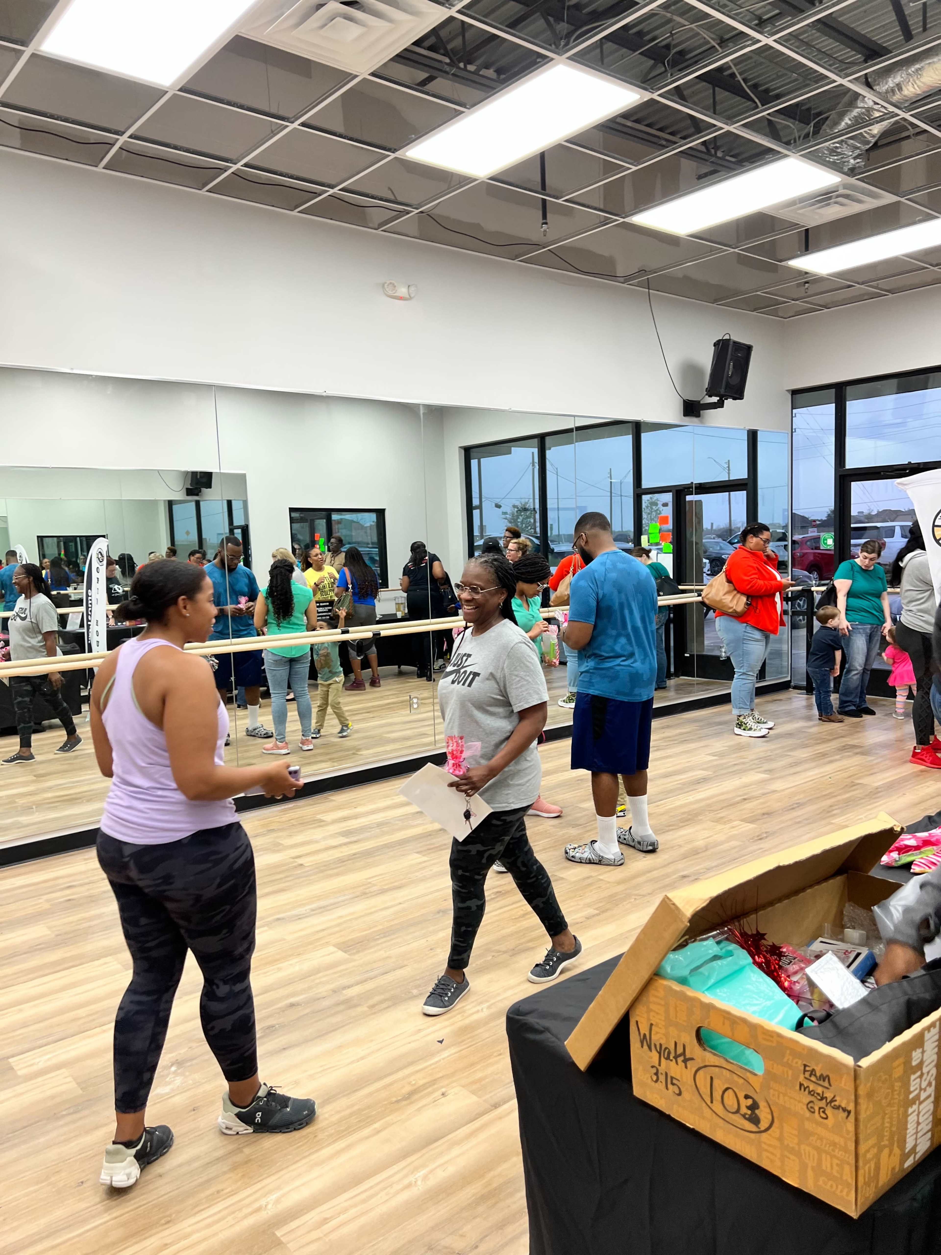 A group of people gather in a well-lit fitness studio with mirrors, some participating in an exercise class and others waiting around, while a box of supplies sits on a table.