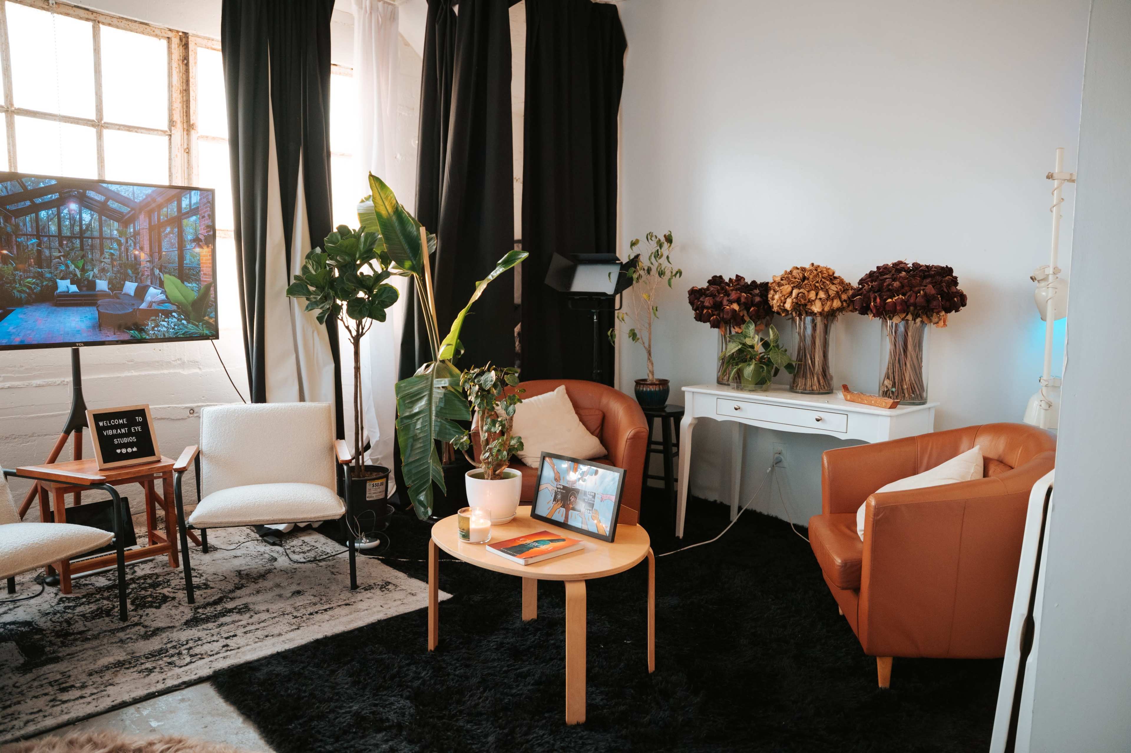 The image shows a cozy interior space featuring two brown armchairs, a round coffee table, a television, various indoor plants, and a side table adorned with dried flowers.