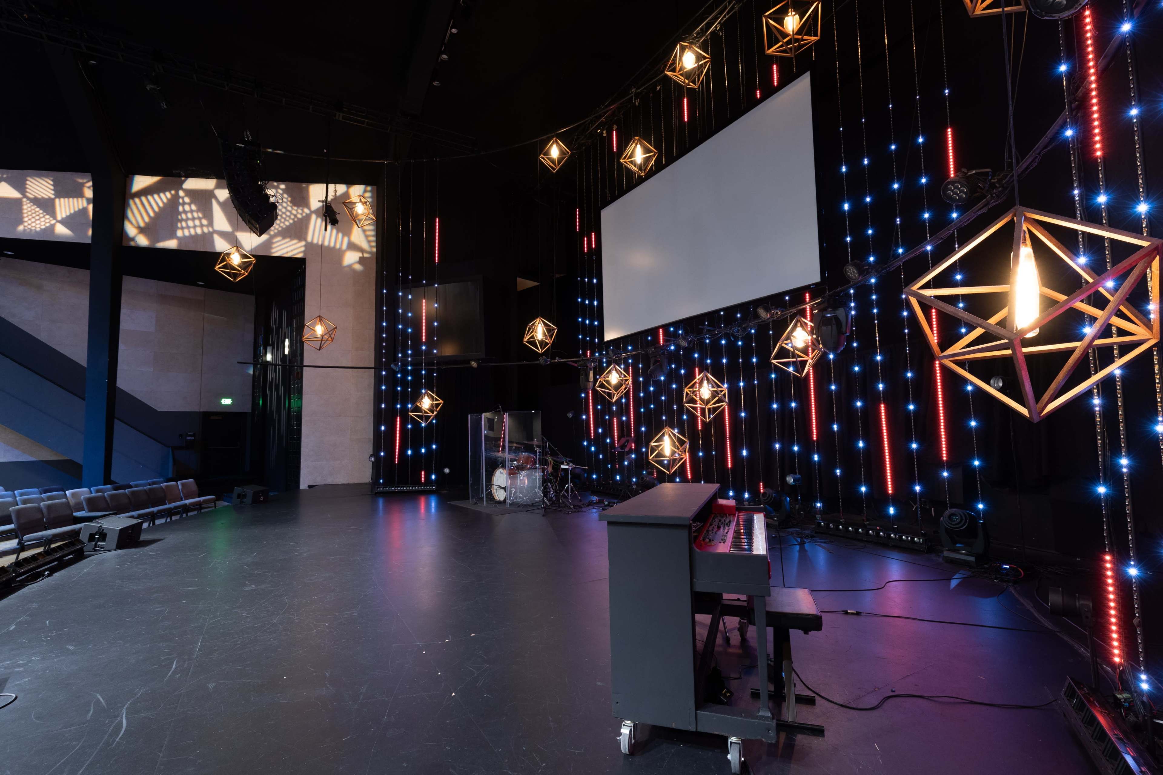 The image shows a modern stage setup with a keyboard, a drum set in the background, and illuminated geometric light fixtures hanging from the ceiling.