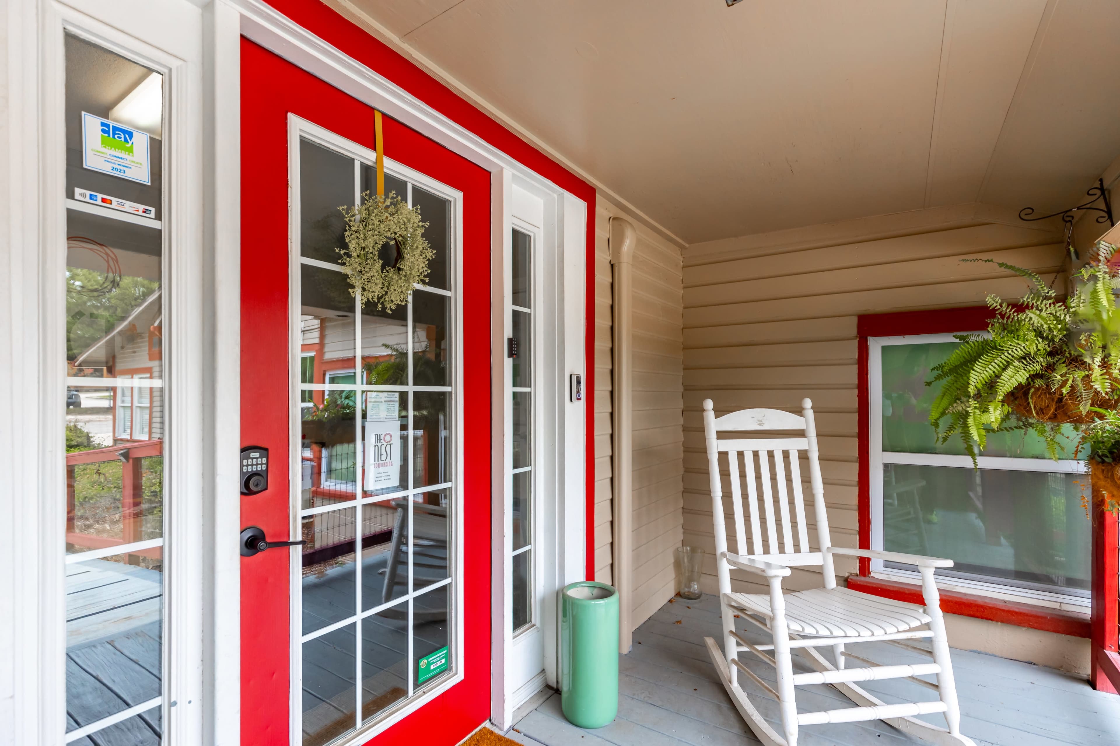 A porch featuring a red front door, a rocking chair, and potted plants, with a wreath hanging on the door.