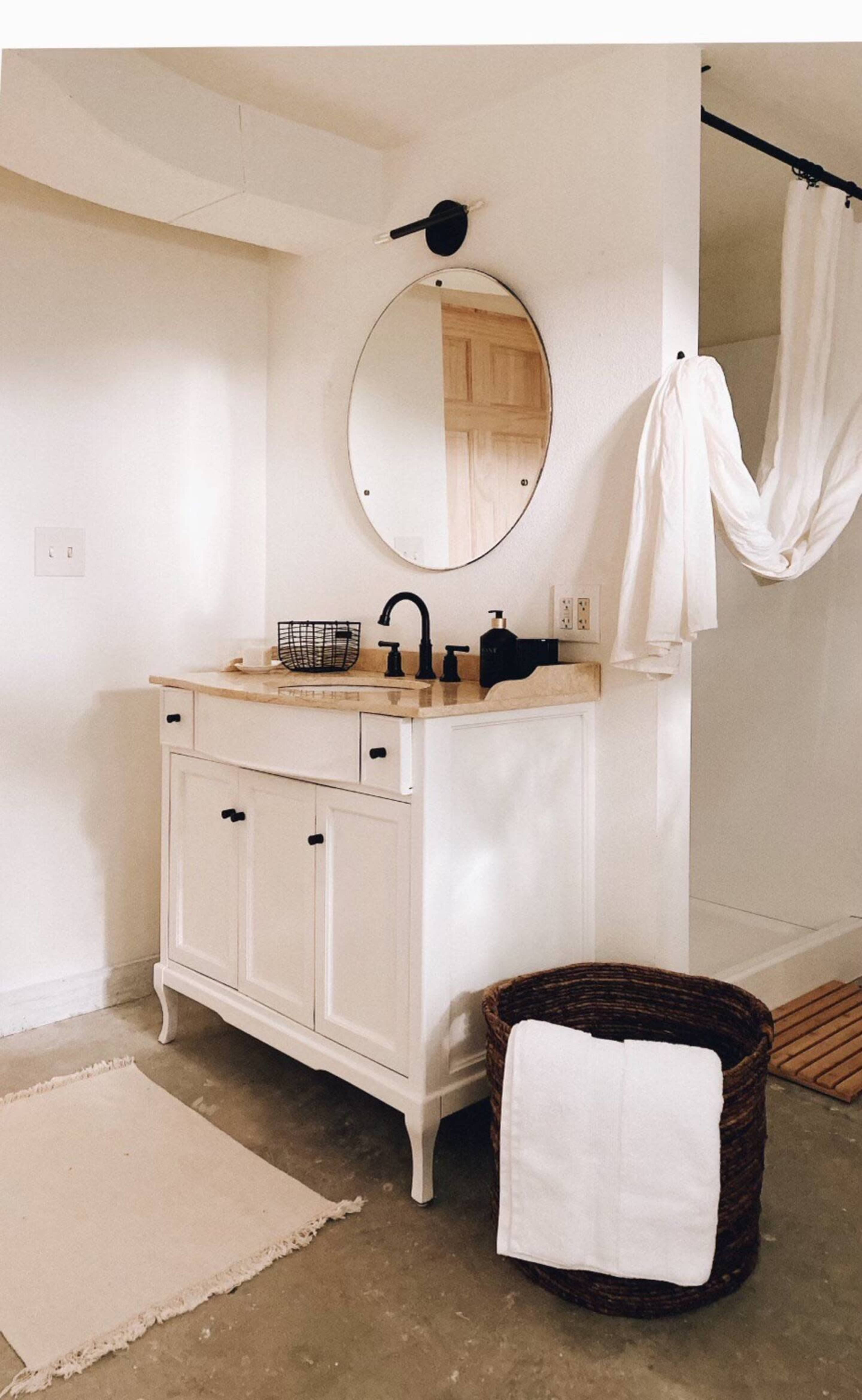 A bathroom featuring a white vanity with a round mirror, a black faucet, and a woven basket alongside a hanging shower curtain.