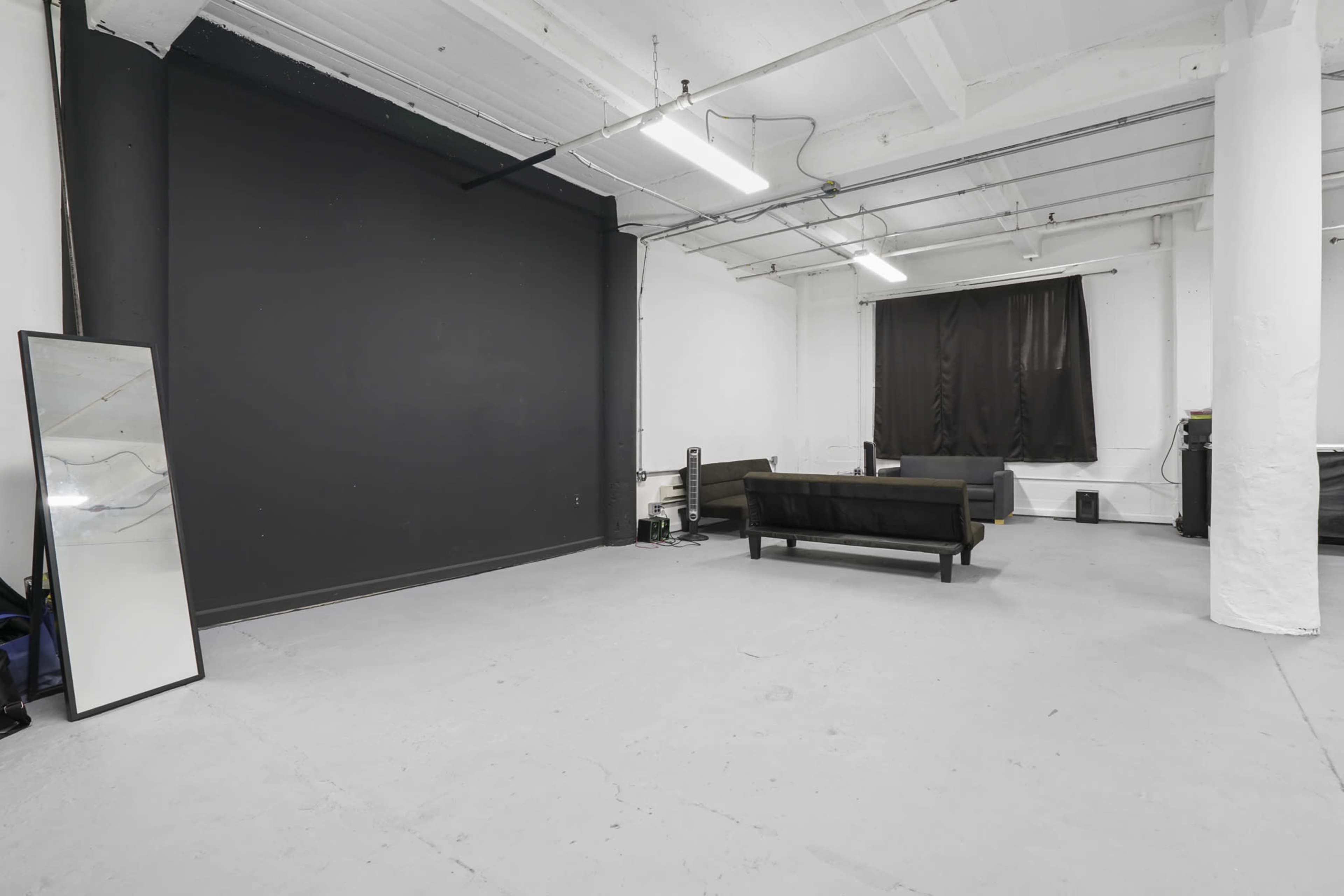 A spacious, empty room features a black wall, a dark curtain on the window, a sofa, and a tall mirror.