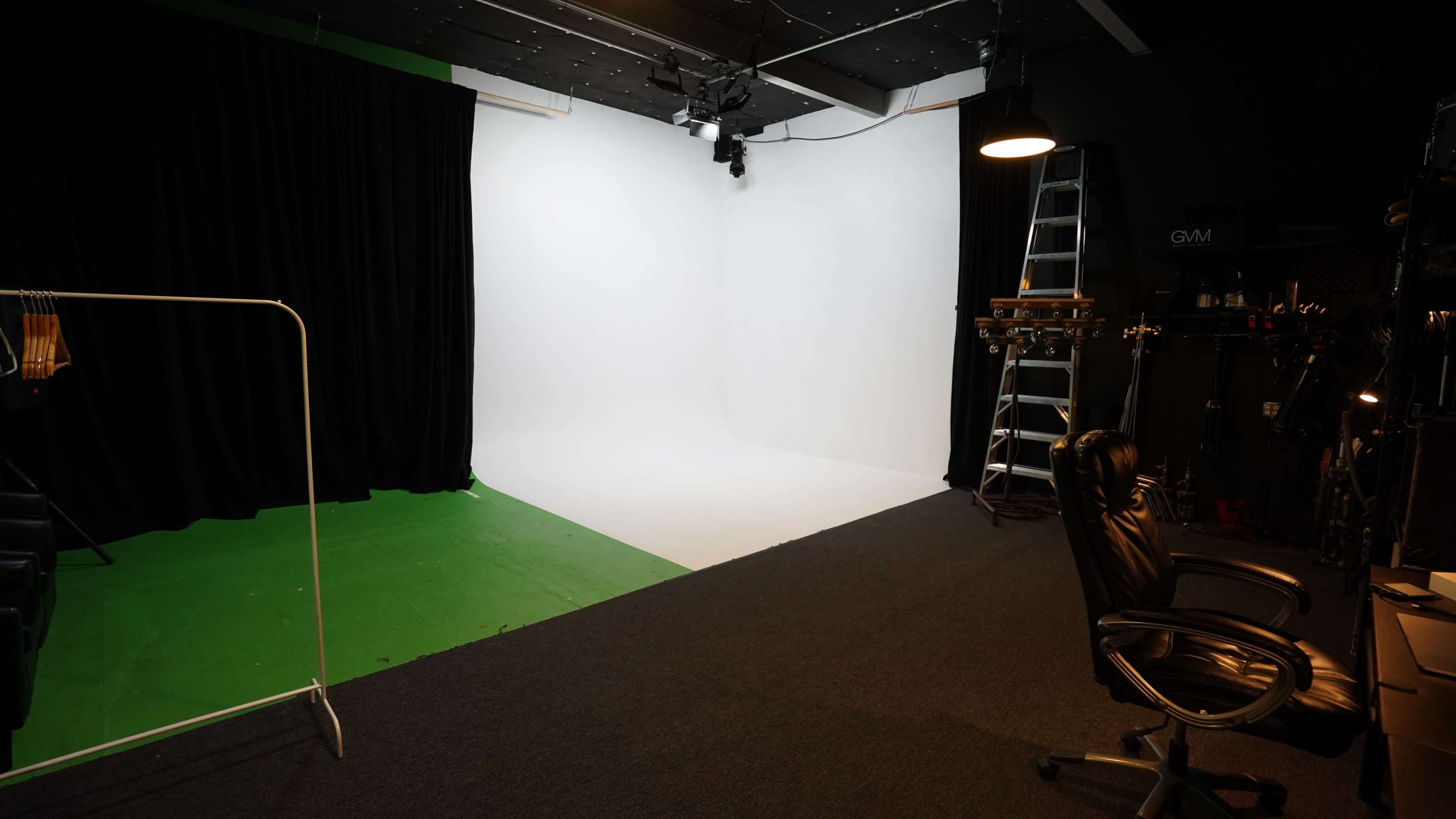 A photography studio with a white backdrop, a black curtain, a green section of the floor, and a black office chair nearby.