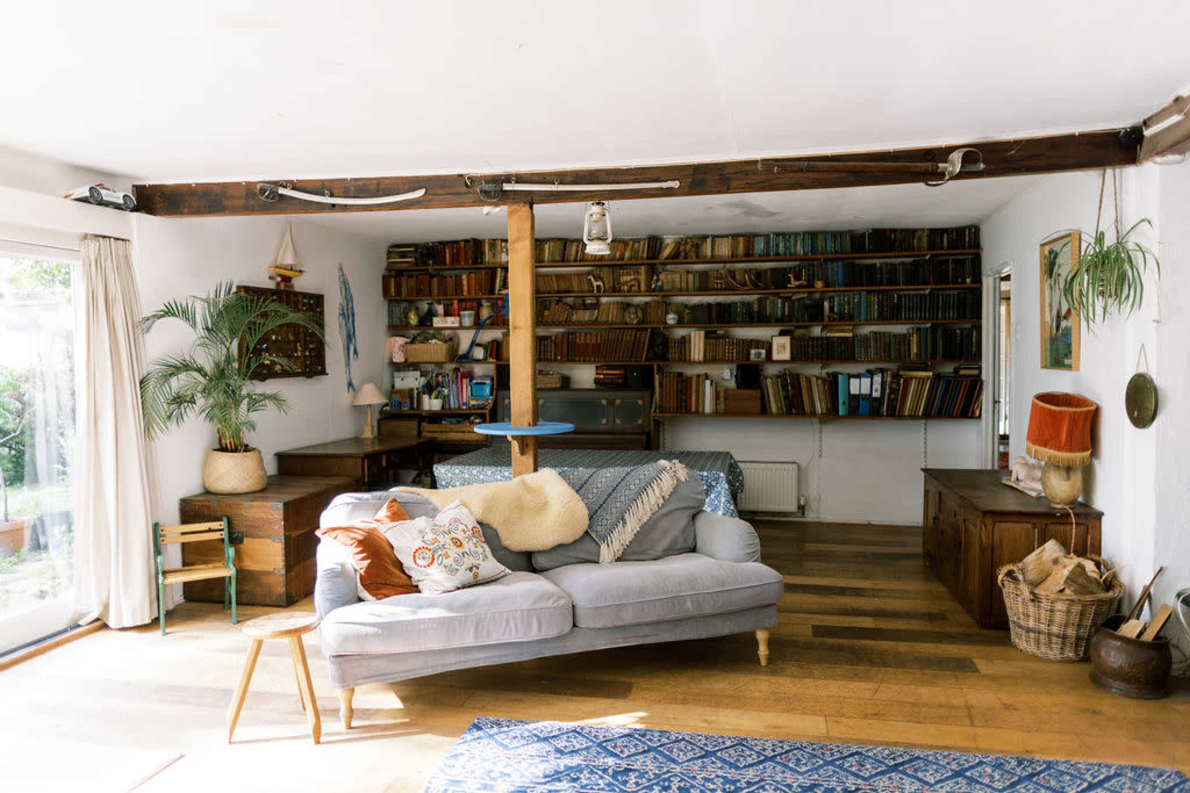 A cozy living room features a sofa with decorative cushions, a bookshelf filled with books, and a wooden beam supporting the ceiling.