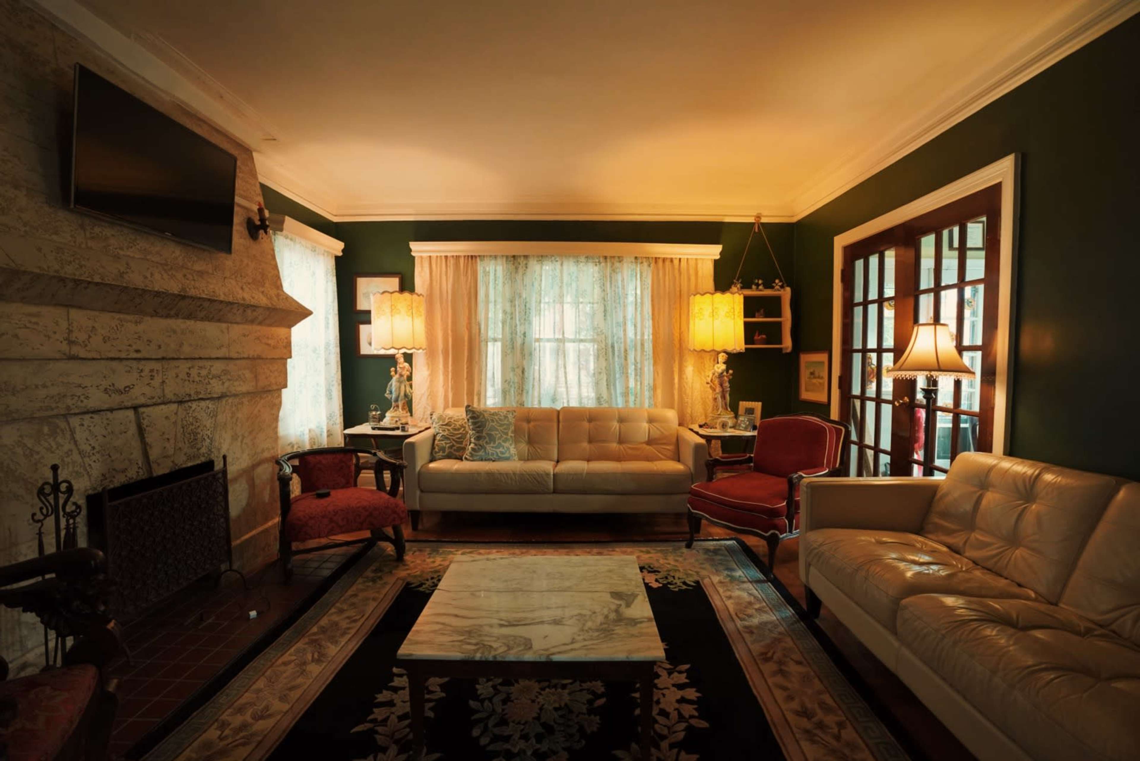 A living room features a beige sofa and cream chairs surrounding a marble coffee table, with green walls and large windows dressed in sheer curtains.