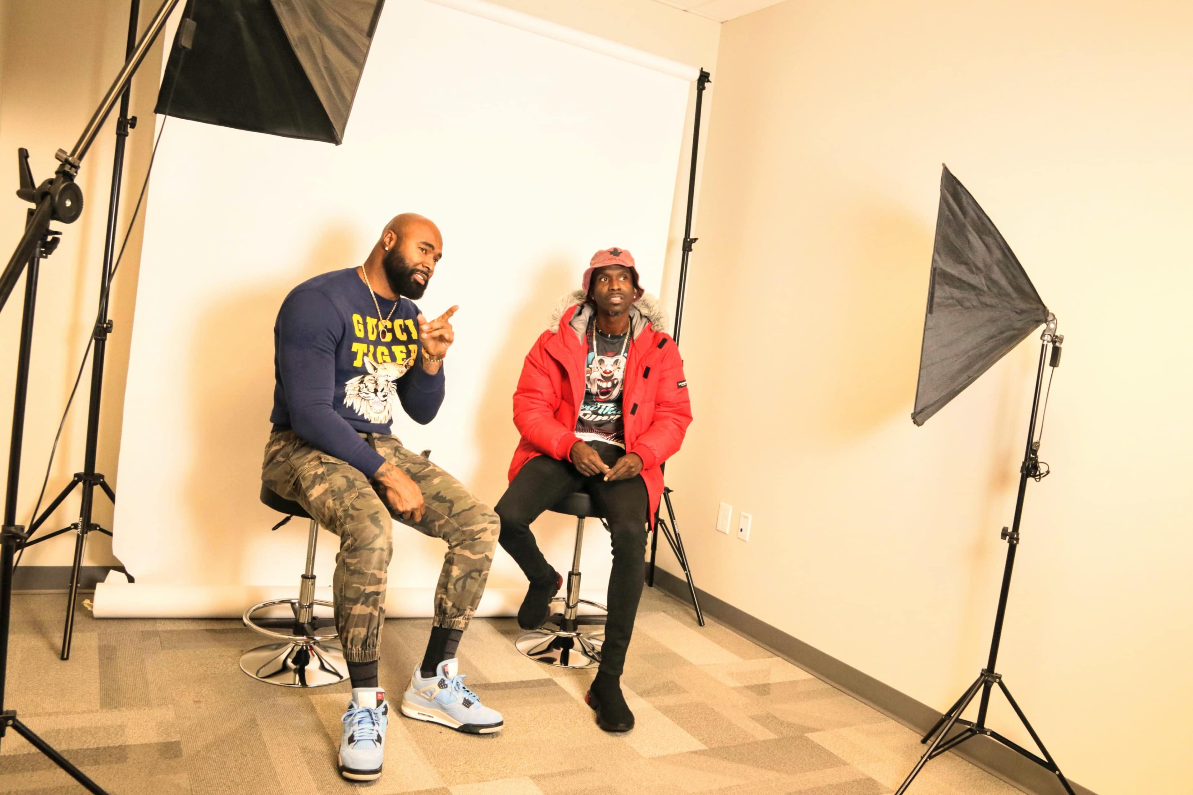 Two men sit on stools in a brightly lit studio, surrounded by photography lighting equipment.
