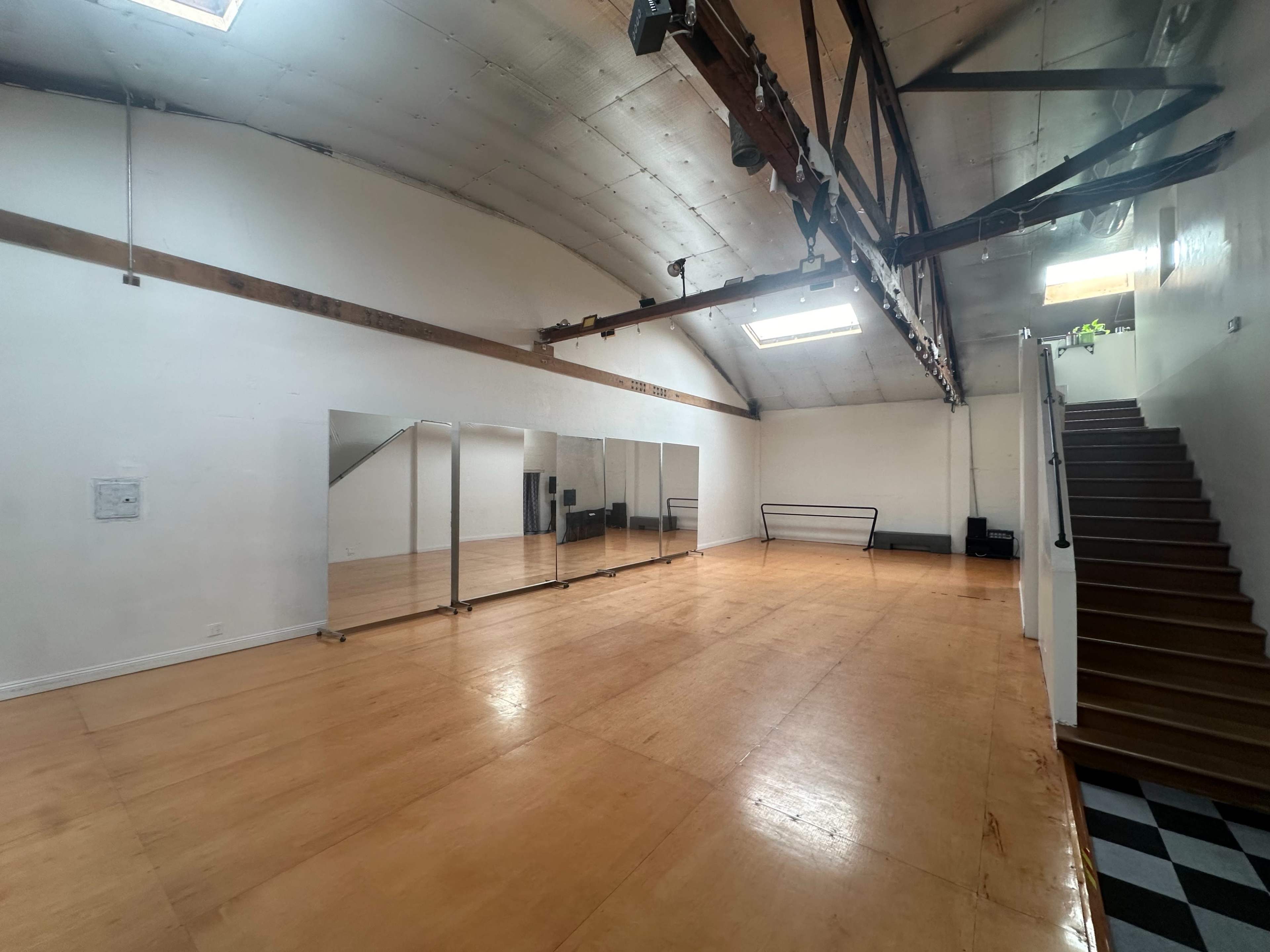 The image shows a spacious, well-lit room with wooden flooring, mirrored walls, and a staircase leading to an upper level.