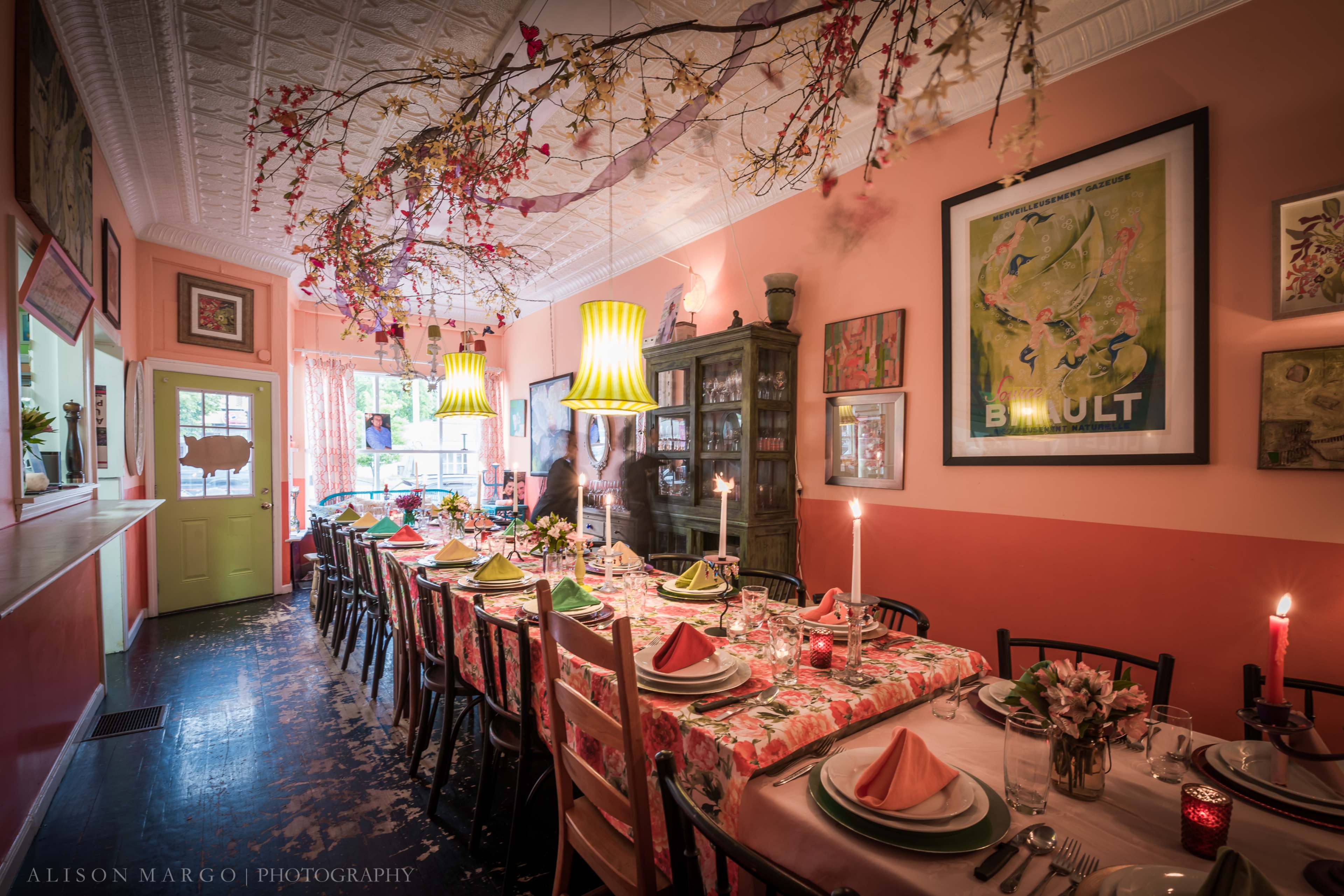 A long dining table is set for a meal in a warmly decorated room, featuring floral tablecloths, vibrant napkins, and soft lighting from hanging lamps.