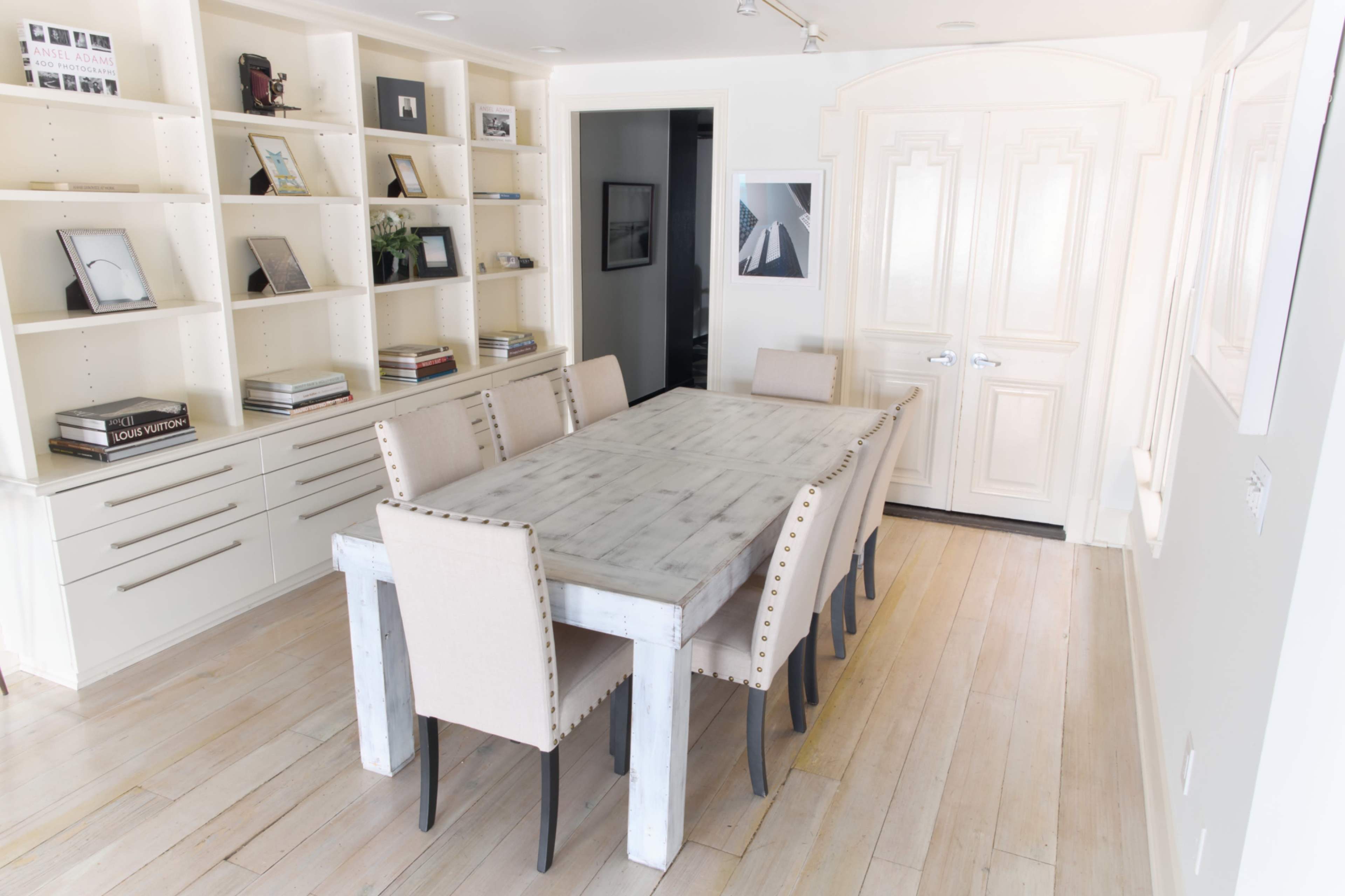 The image shows a modern dining area featuring a long rectangular wooden table surrounded by beige chairs, with built-in shelves and a door in the background.