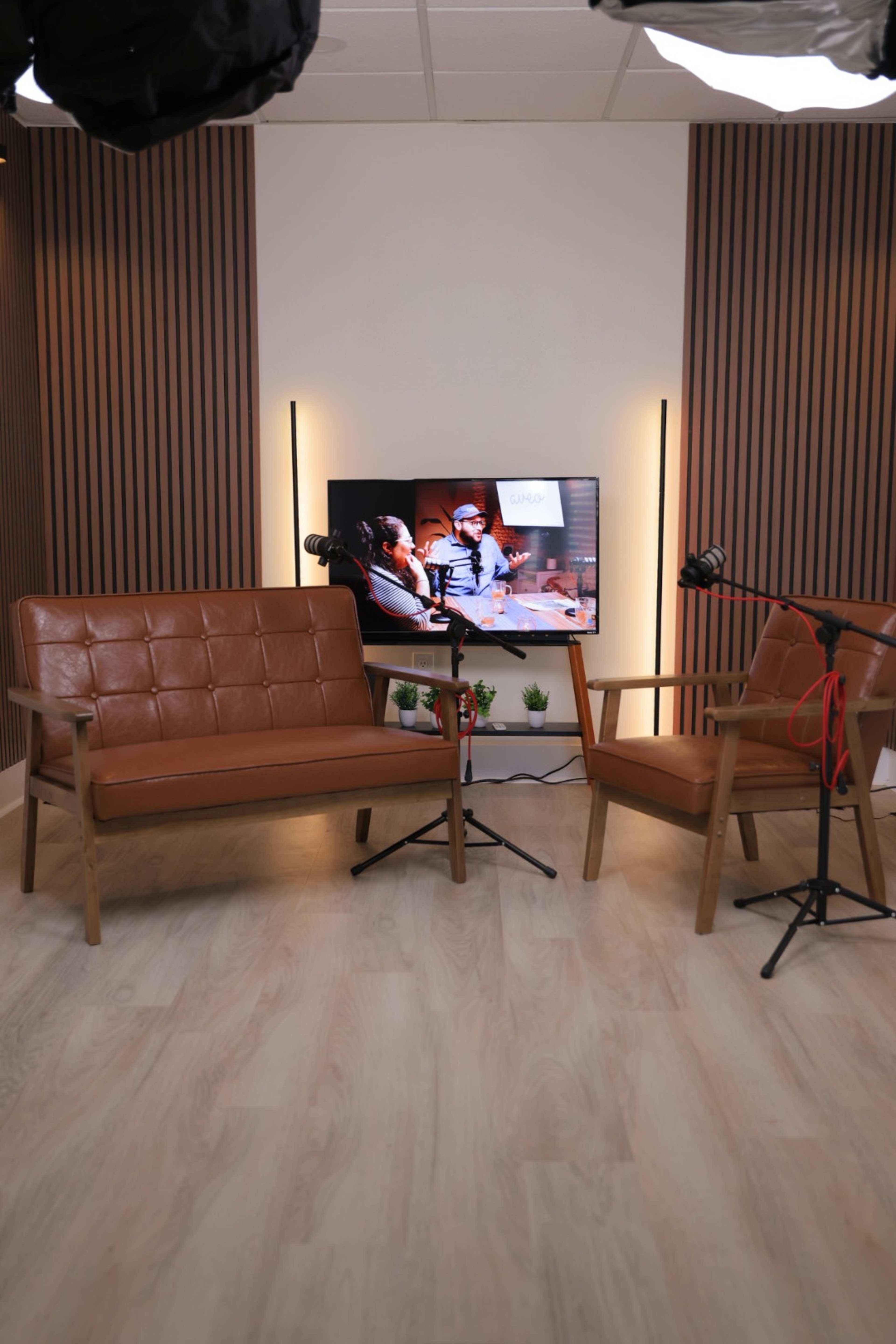 The image shows a minimalist recording setup featuring two leather chairs and a television displaying a show, with soft lighting and wooden wall panels in the background.