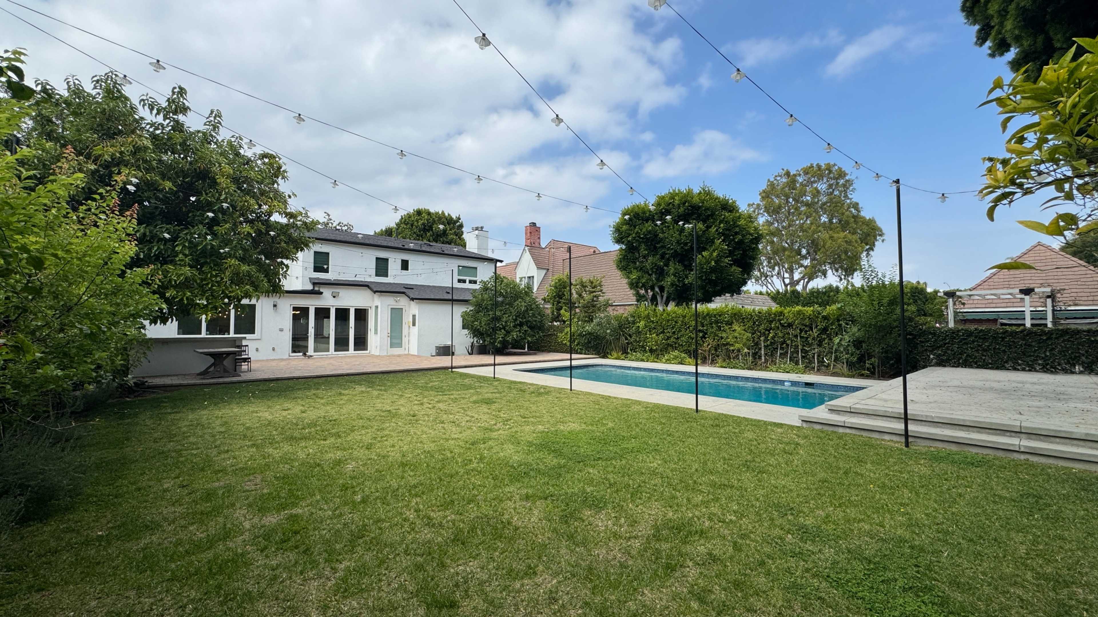 The image shows a backyard with a swimming pool, surrounded by greenery, a paved area, and a white house in the background under a partly cloudy sky.