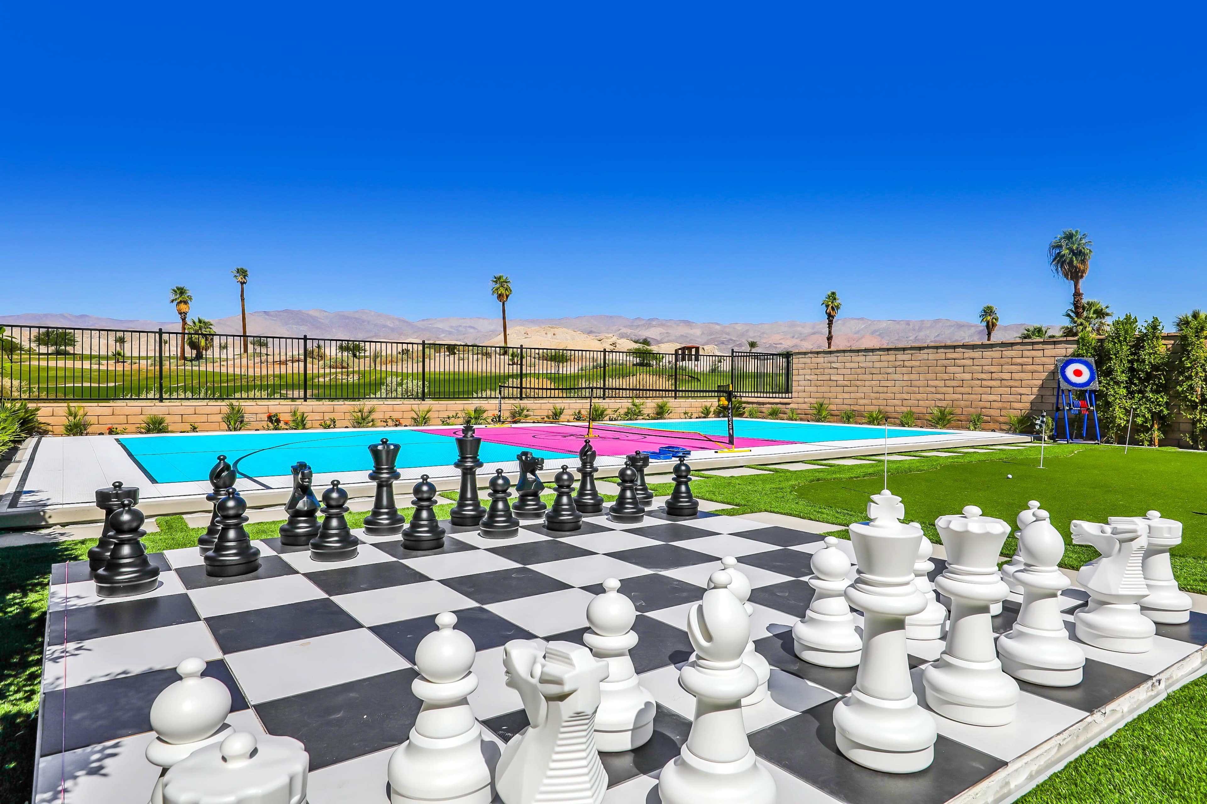 A large outdoor chess set is positioned on a checkerboard surface near a swimming pool, with mountains in the background and a clear blue sky above.