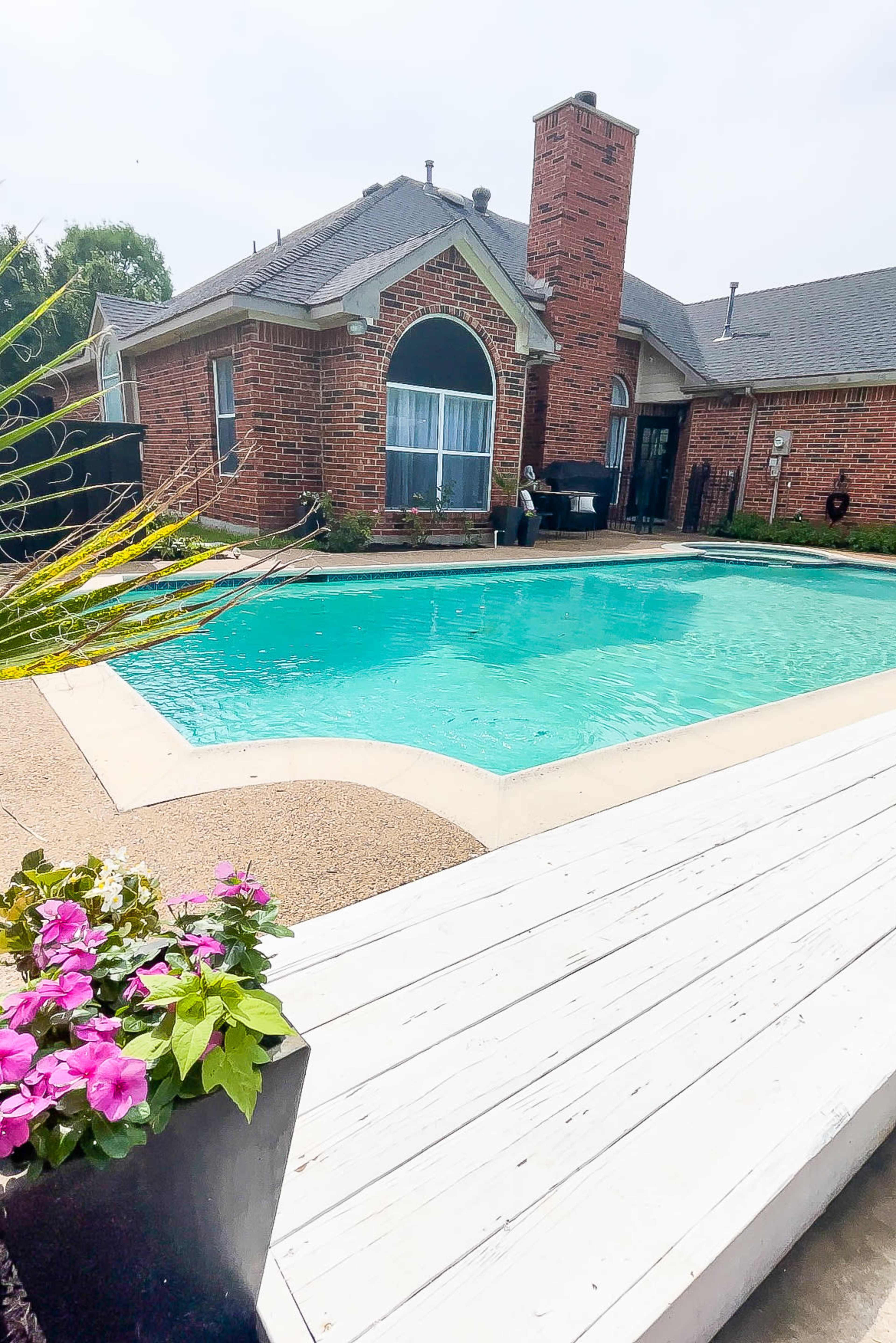 A brick house with a chimney overlooks a clear swimming pool surrounded by a concrete deck and flower pots.