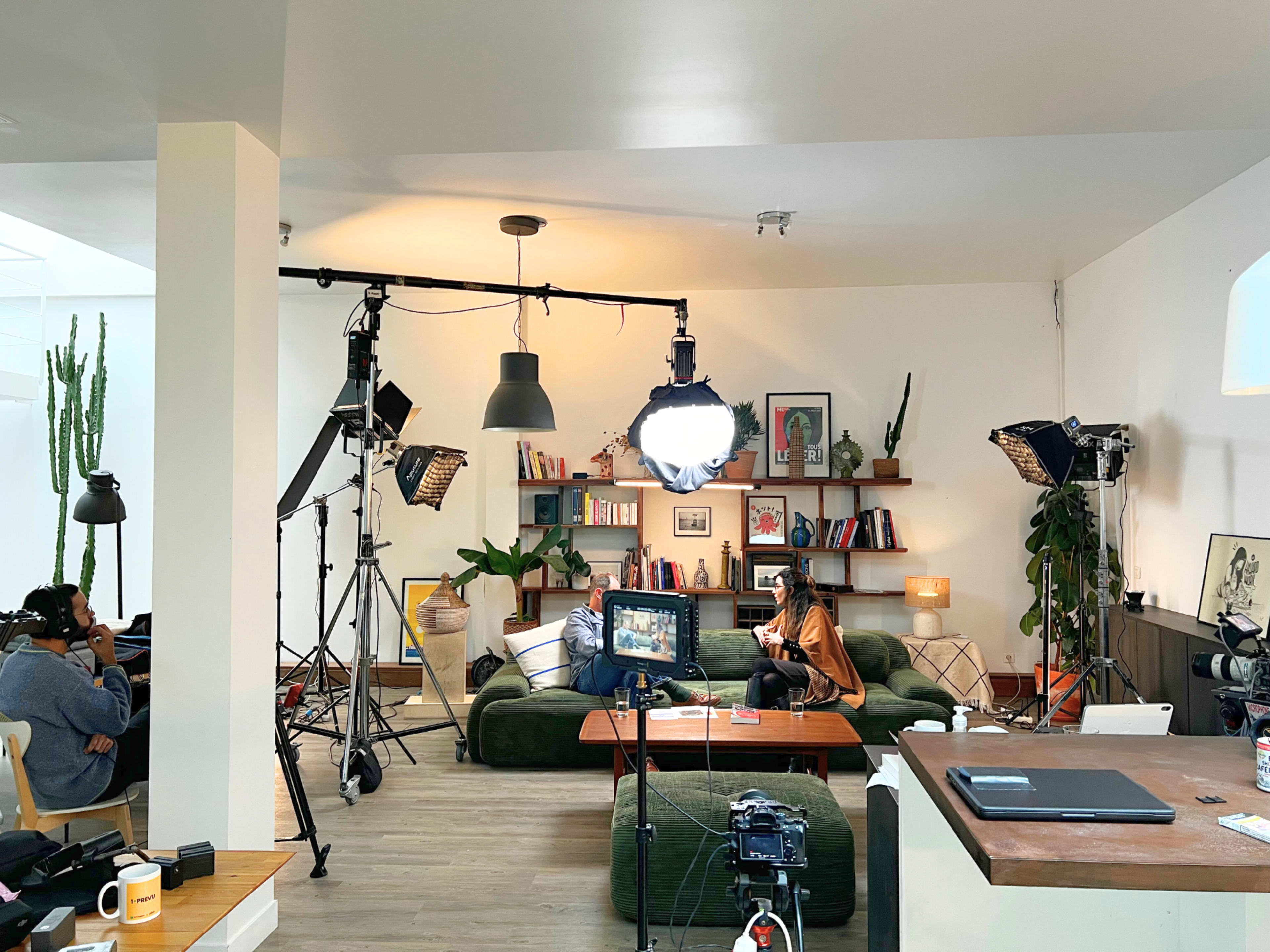 The image shows a cozy studio space set up for filming, featuring cameras, lighting equipment, and a seating area with bookshelves and plants in the background.