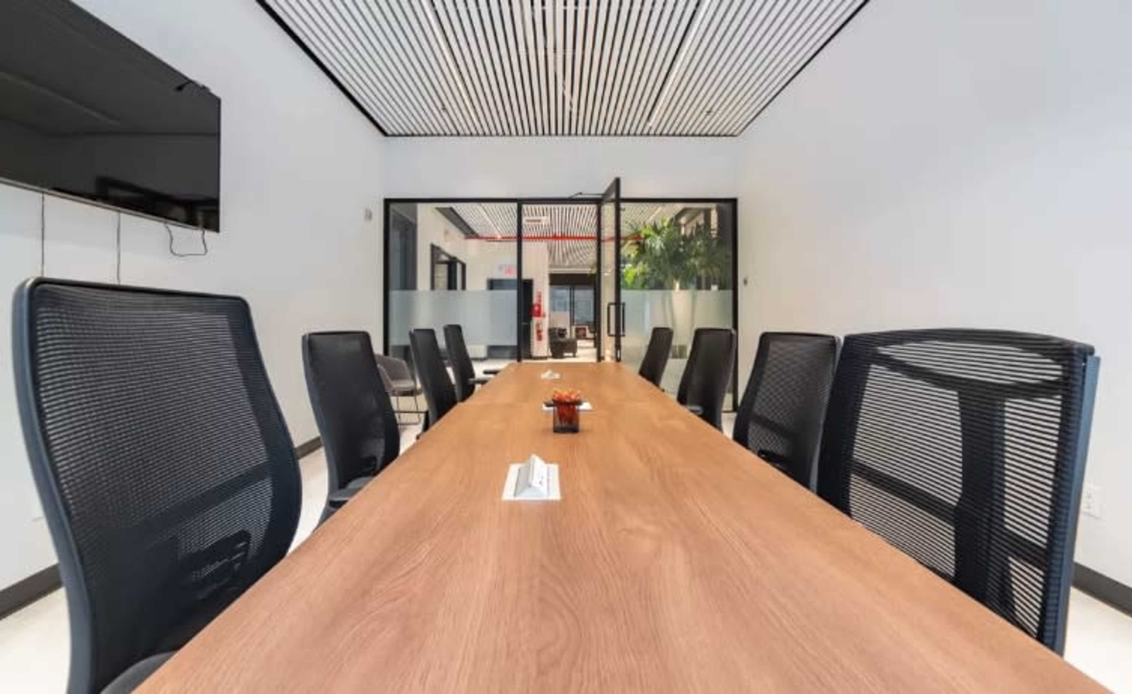 A long wooden conference table is surrounded by black mesh chairs in a modern meeting room with glass walls.