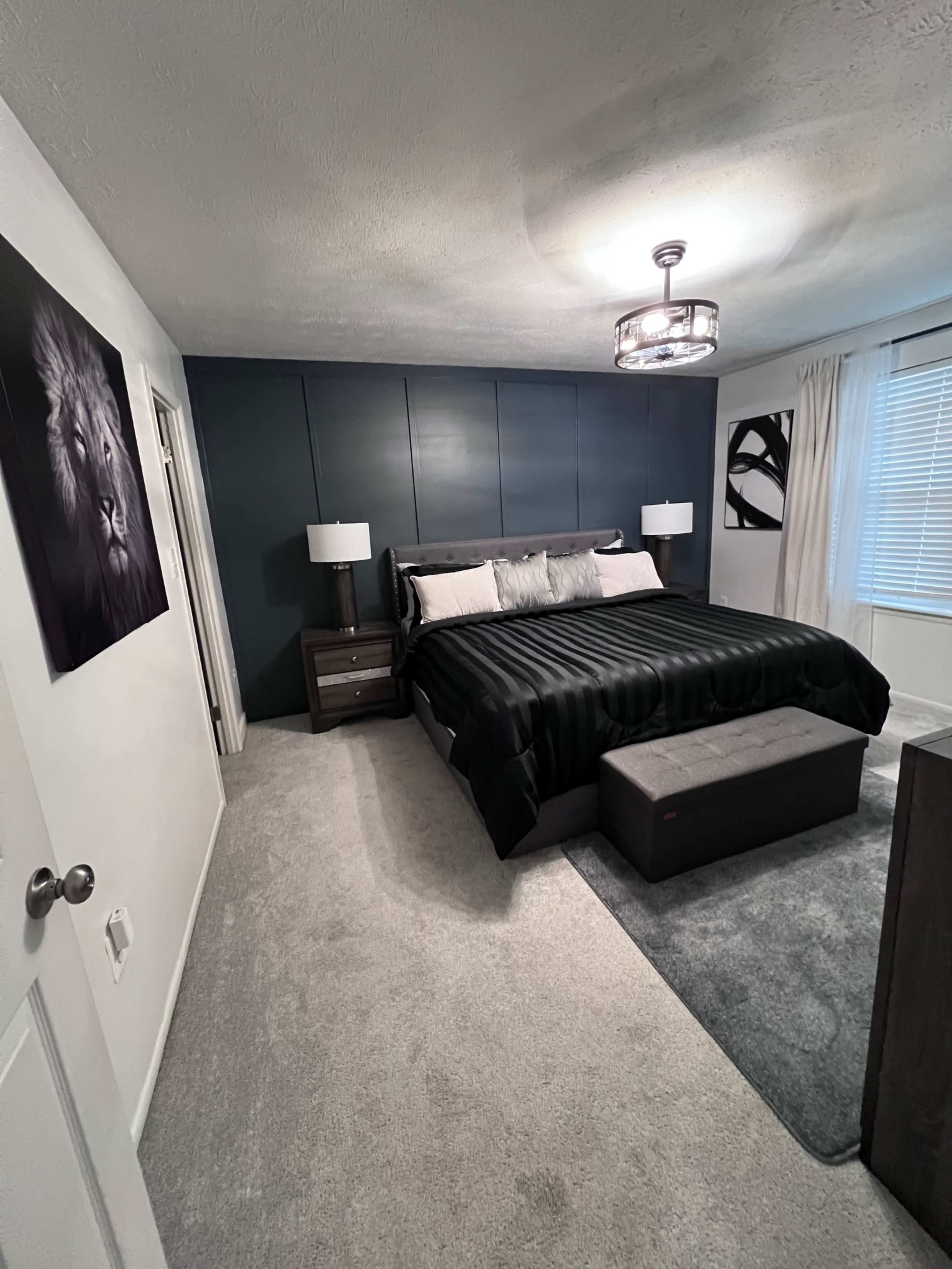 The image shows a bedroom featuring a large bed with black bedding, two bedside lamps, a gray upholstered bench at the foot of the bed, and a lion portrait on the wall.