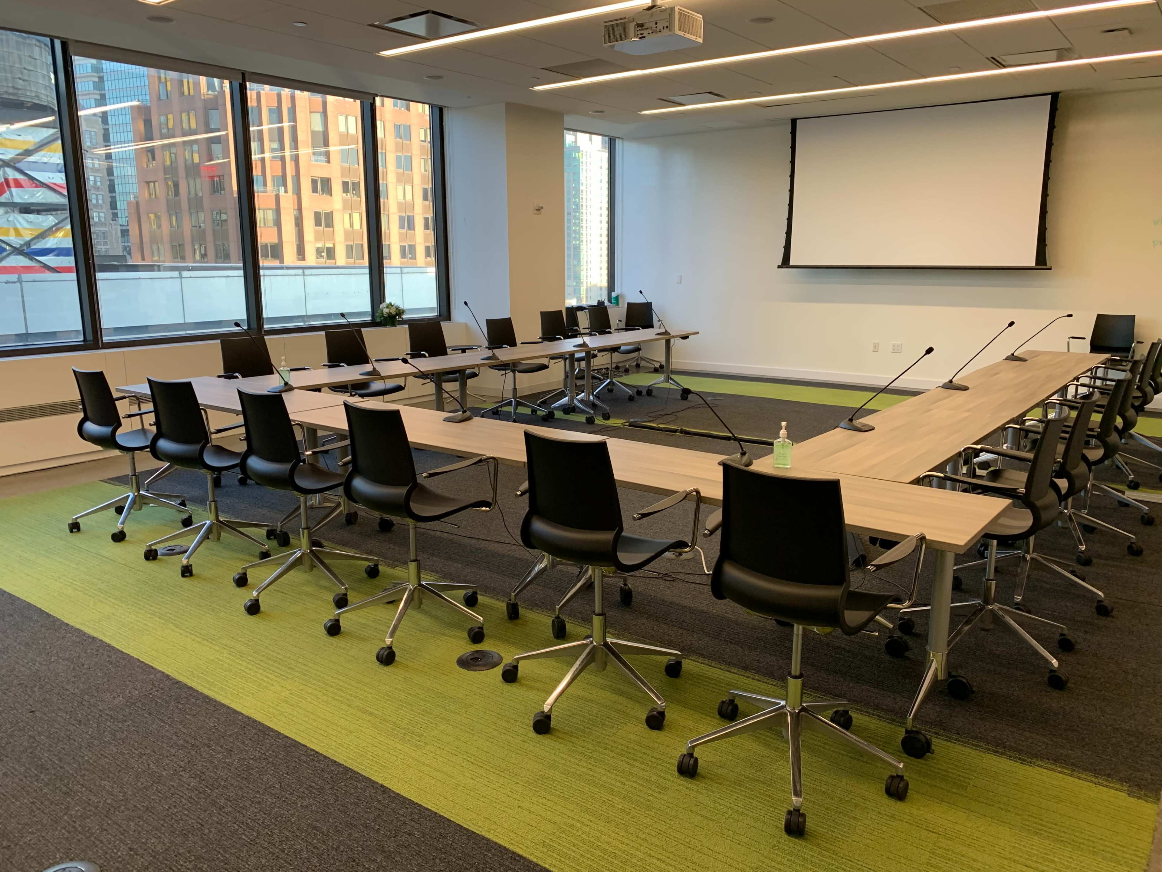 A large conference room with a long table surrounded by office chairs, large windows revealing a cityscape, and a projector screen mounted on the wall.