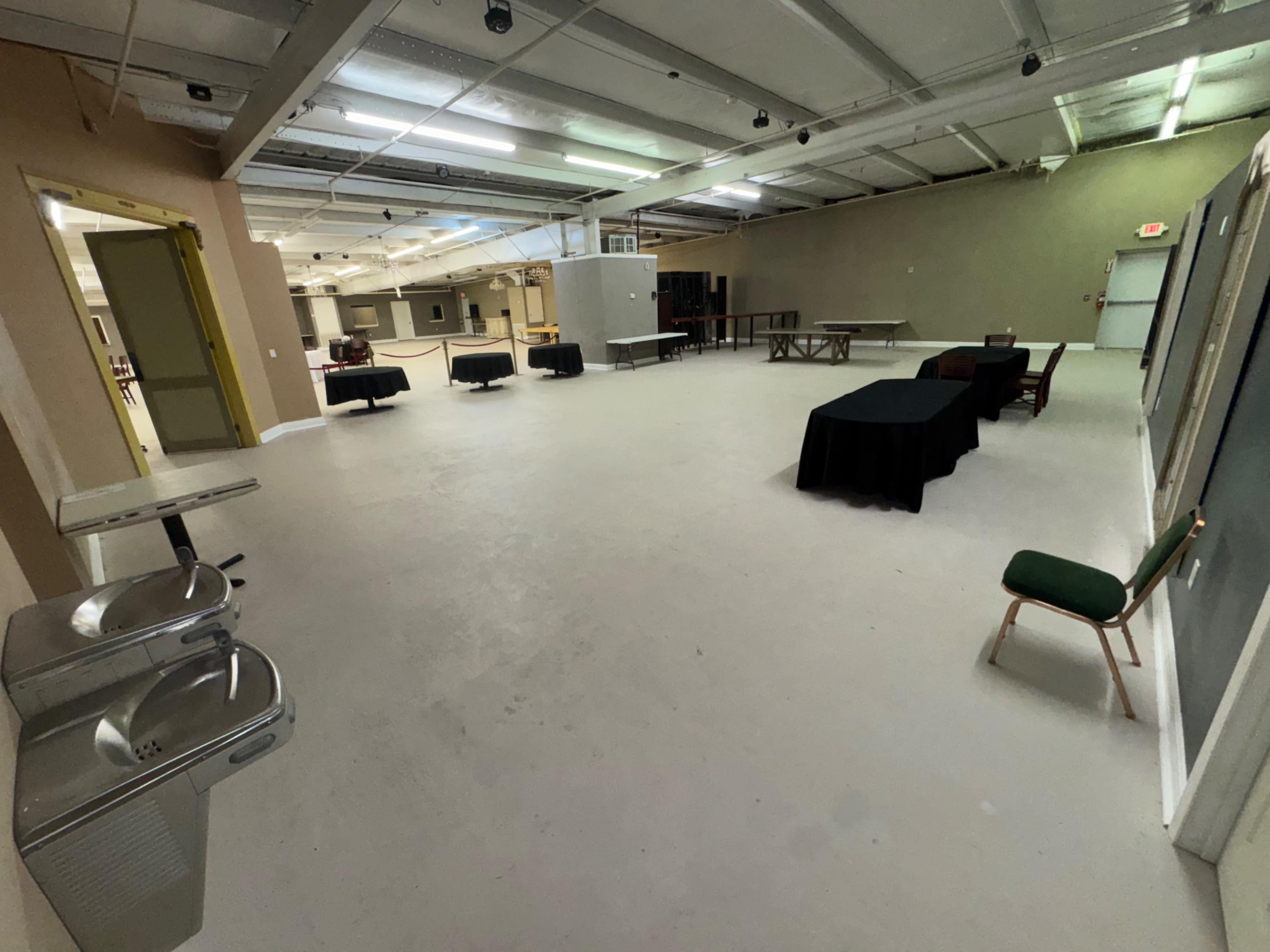 The image shows a large, empty indoor space with a few tables set up around the perimeter and a water fountain against one wall.