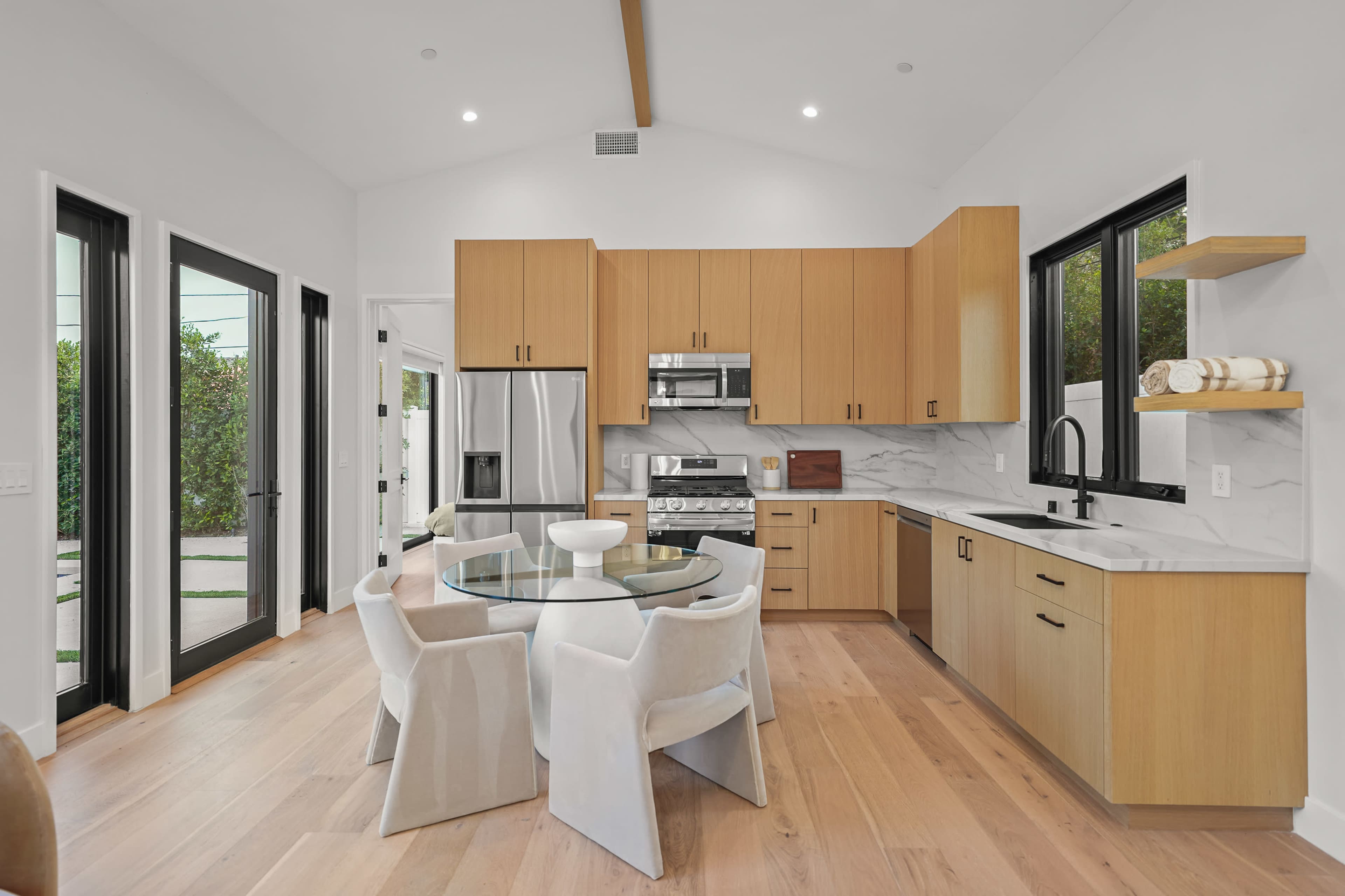 A modern kitchen features wooden cabinetry, stainless steel appliances, a glass dining table, and large windows that provide natural light.