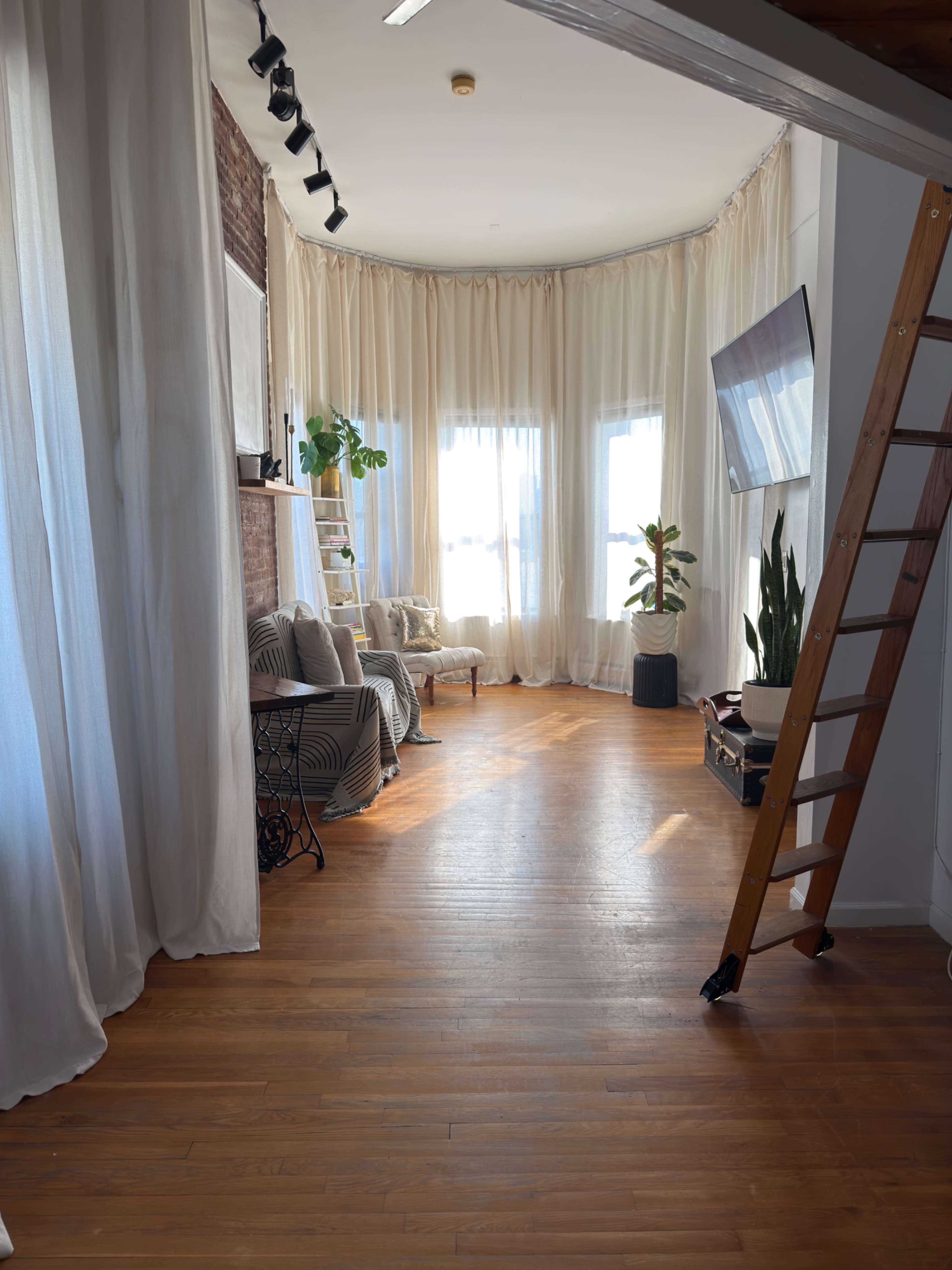 The image shows a bright, airy room with large windows, wooden floors, a cozy seating area, and decorative plants, framed by sheer curtains.