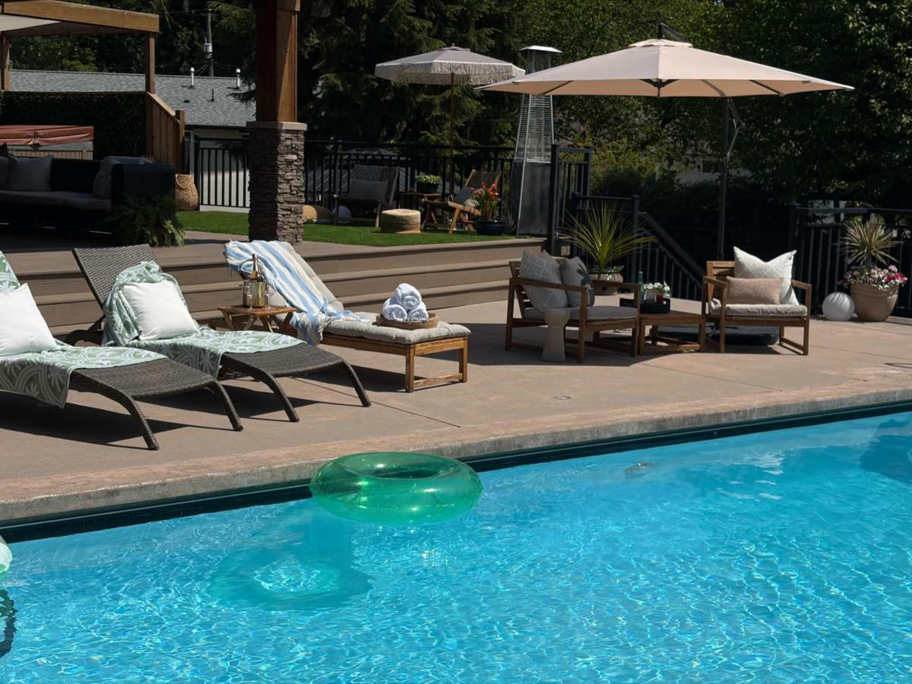 The image shows a swimming pool surrounded by lounge chairs, a coffee table, and an umbrella, with a green inflatable ring floating in the water.