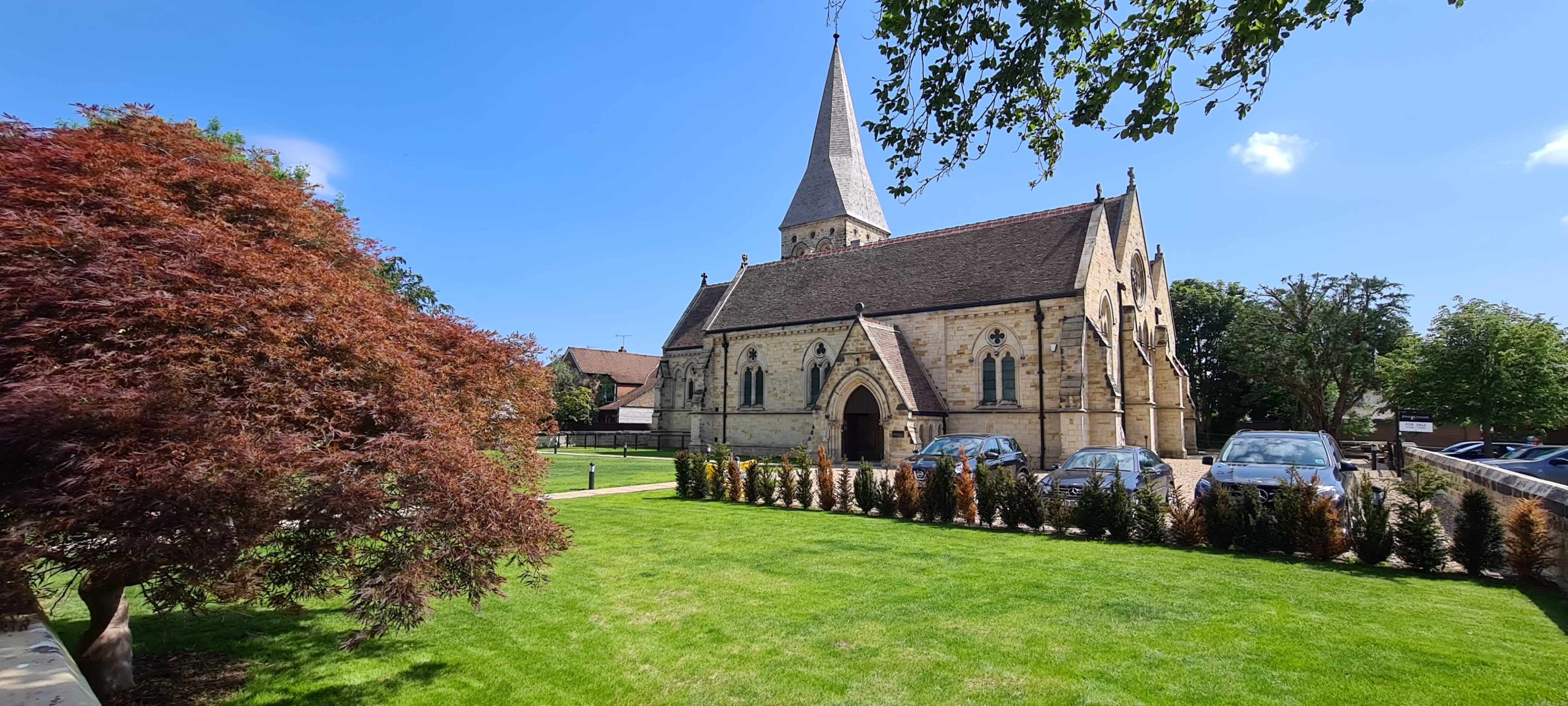 A stone church with a tall spire is surrounded by landscaped grass and trees on a clear day.