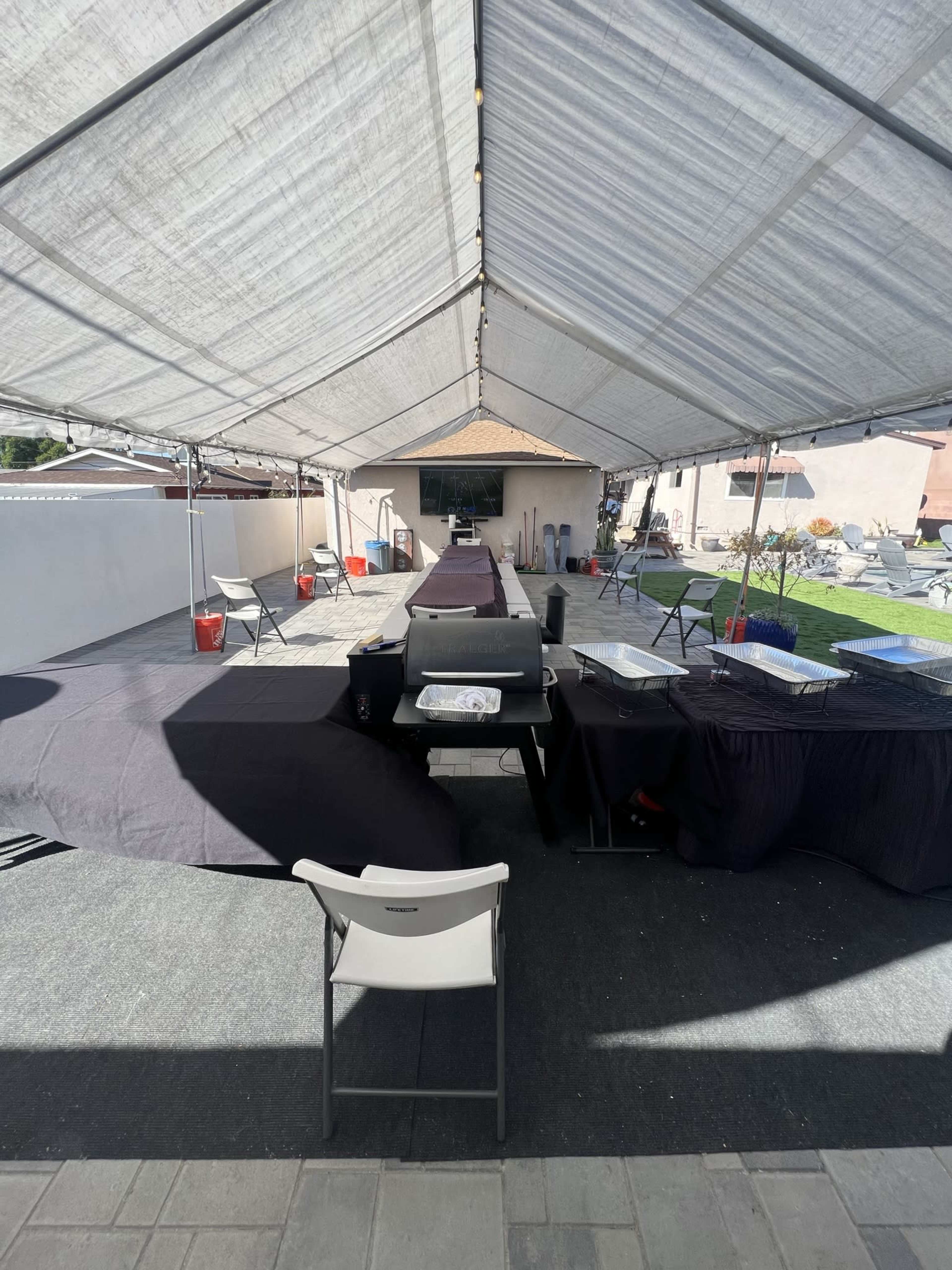 A long table is covered with black cloths beneath a large tent, with empty chairs and a barbecue grill positioned at one end.