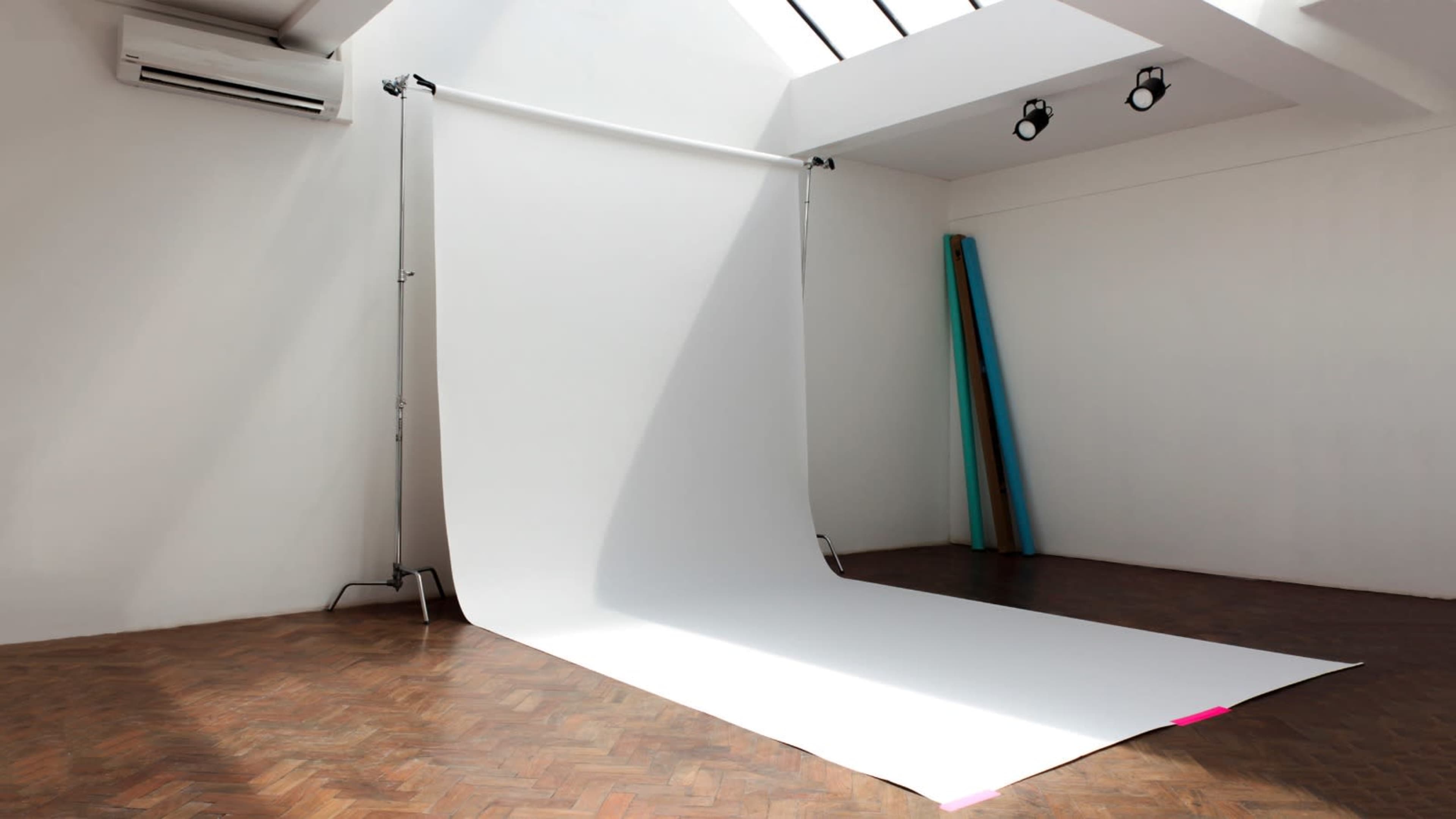 A photography studio featuring a large white backdrop on a wooden floor, with colorful rolls of paper leaning against the wall.