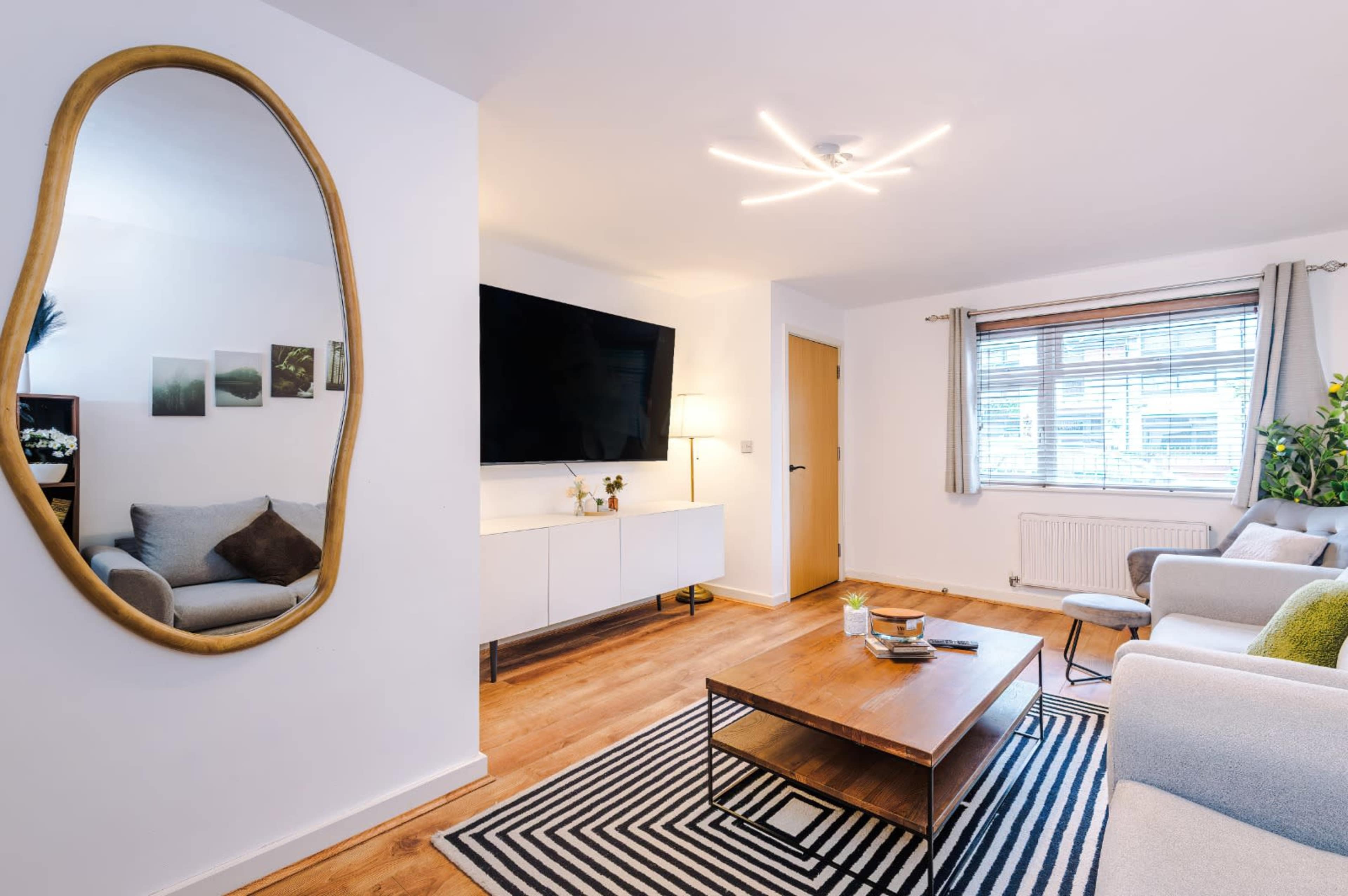 The image shows a modern living room featuring a large wall-mounted television, a rectangular coffee table on a striped rug, and two couches flanking the space, while a stylish mirror hangs on the wall.