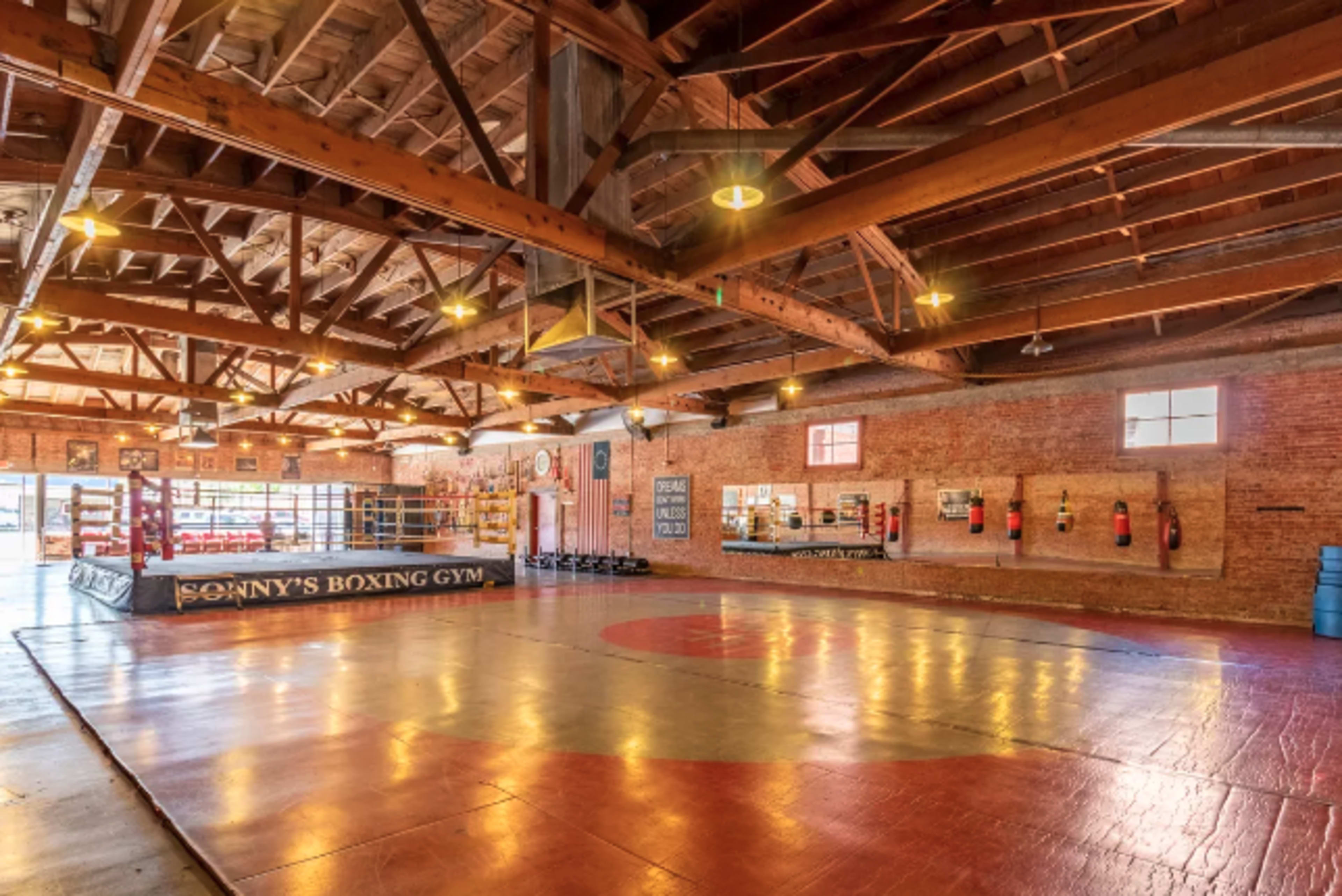 The image shows an interior view of a boxing gym with a large training area, boxing ring, and various equipment along the walls.
