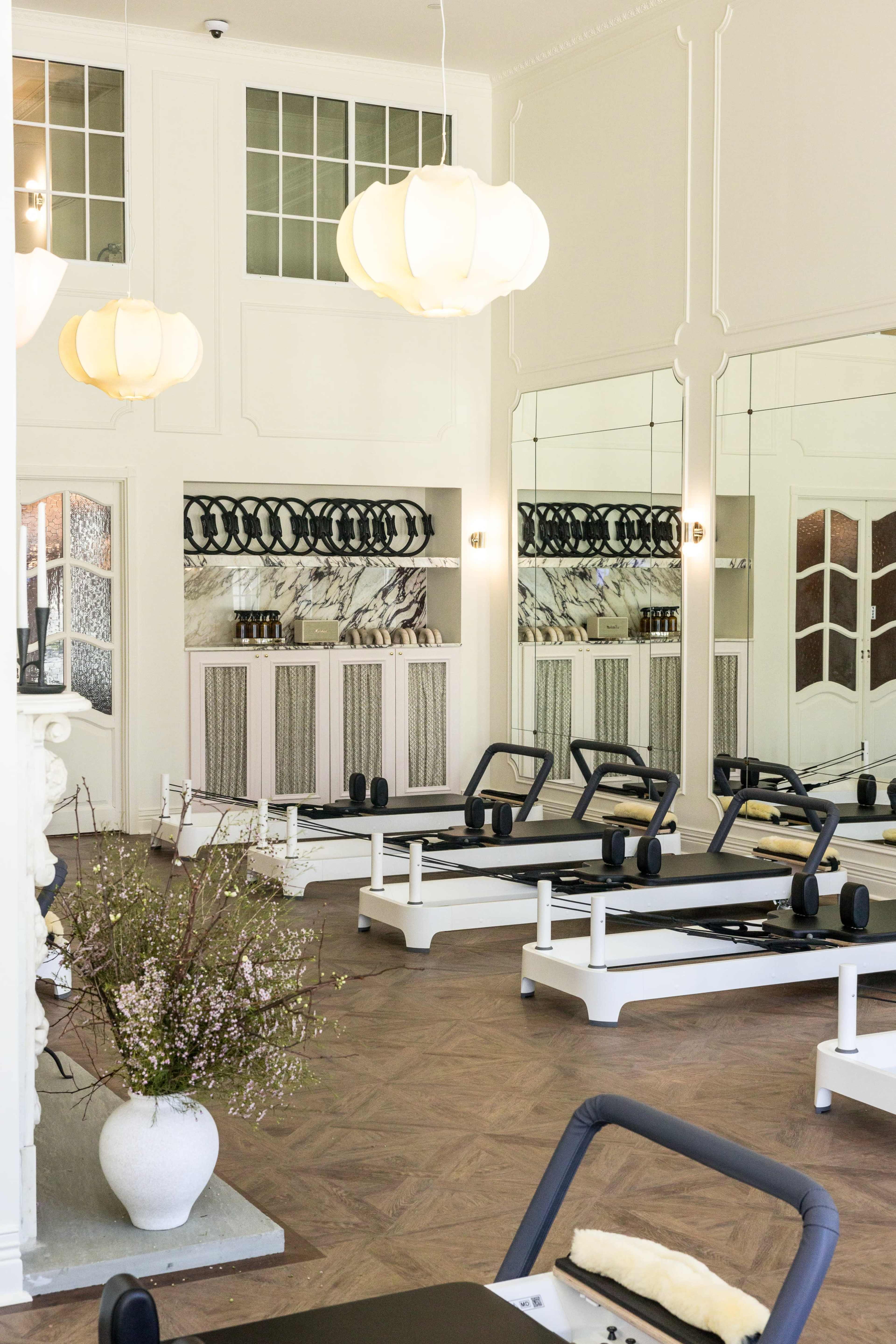 The image features a spacious and well-lit pilates studio with reformer machines arranged neatly, large mirrors on the walls, and elegant light fixtures hanging from the ceiling.