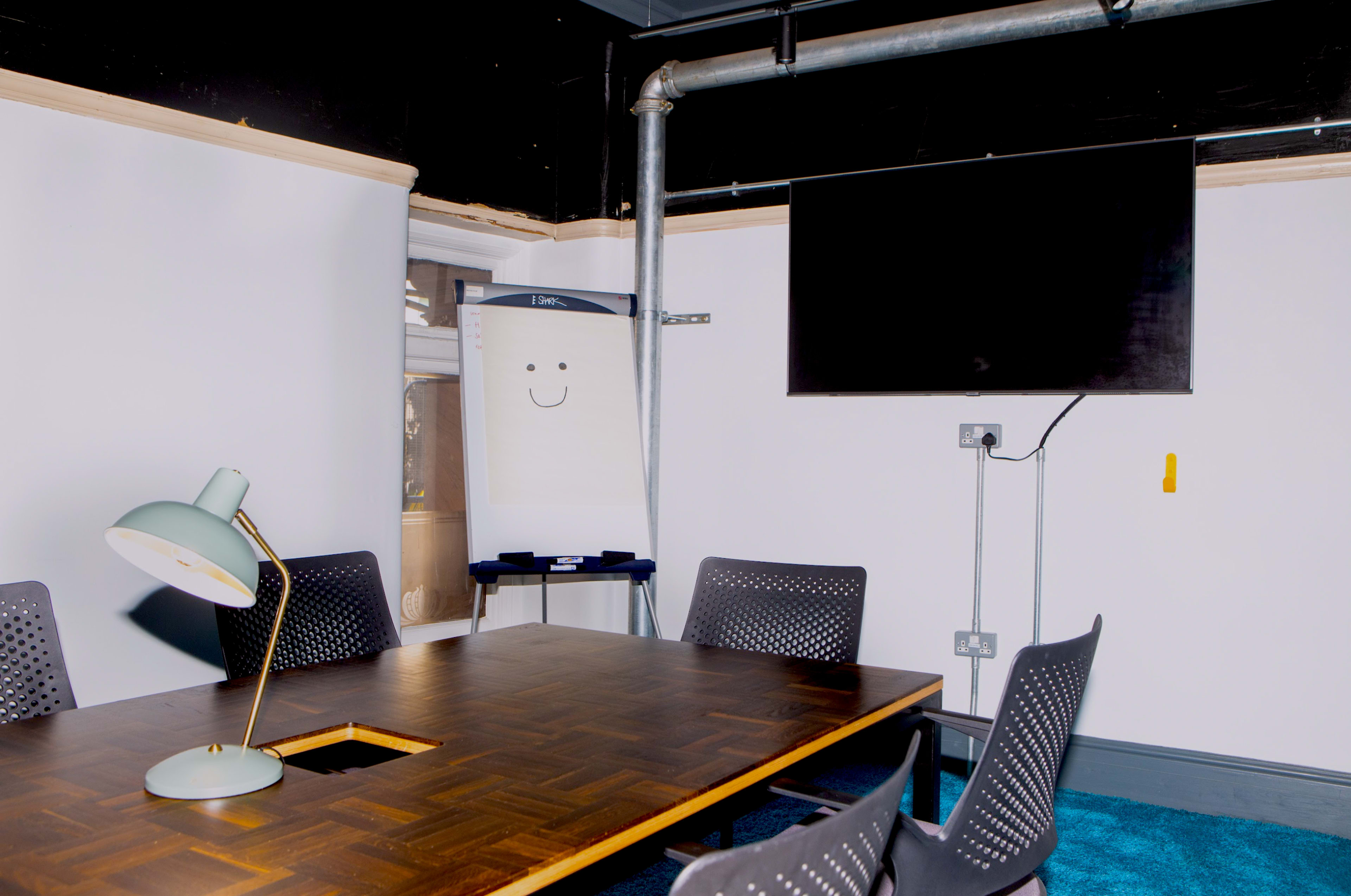 A conference room features a wooden table surrounded by black chairs, with a whiteboard displaying a smiley face and a television mounted on the wall.