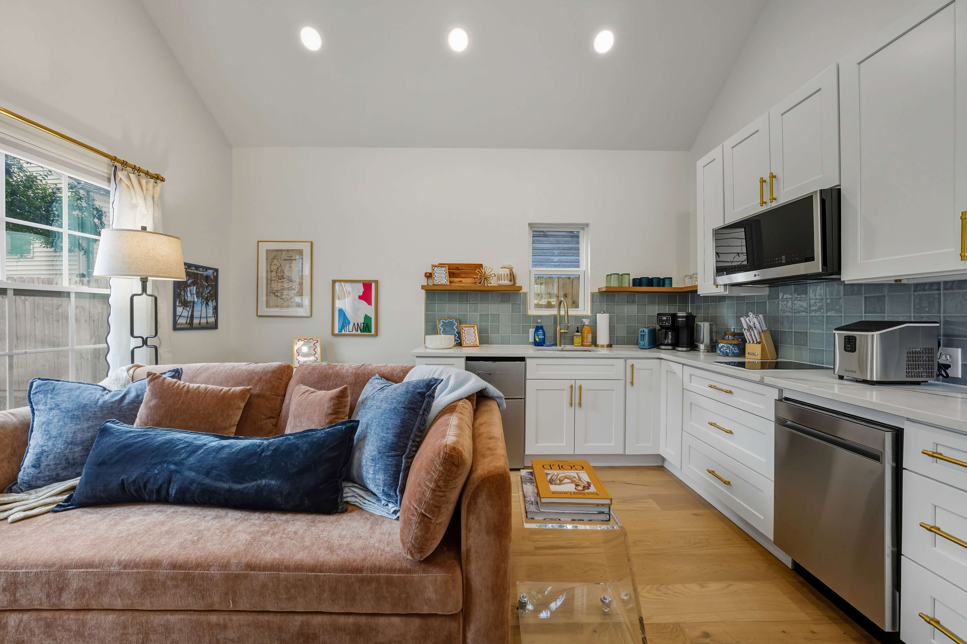 A cozy living area features a plush, beige couch adorned with blue pillows, alongside a modern kitchenette equipped with stainless steel appliances and light green tile backsplash.