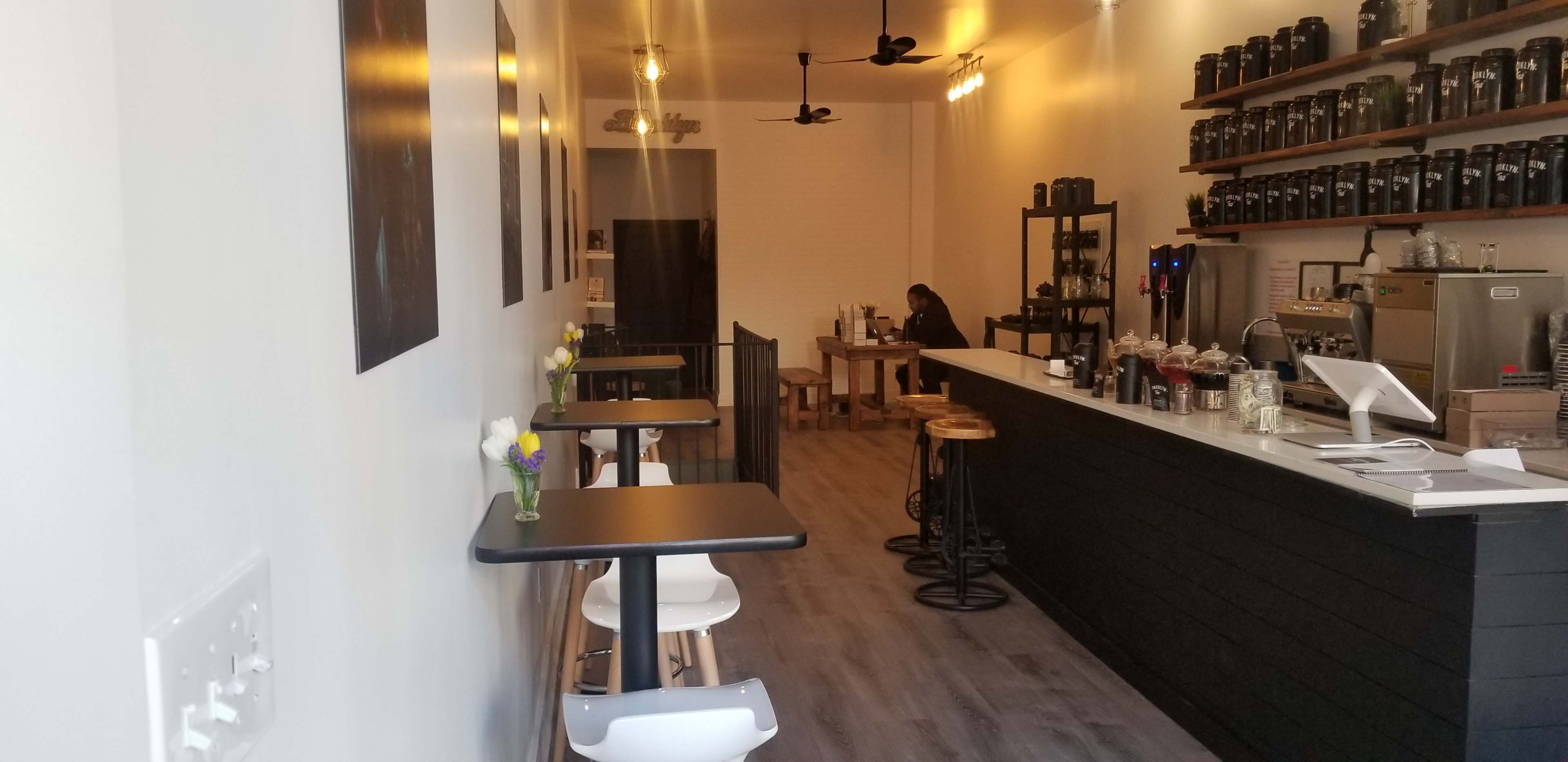 The image shows a modern, minimalist coffee shop with a barista station, tables along the walls, and a customer seated in the background.