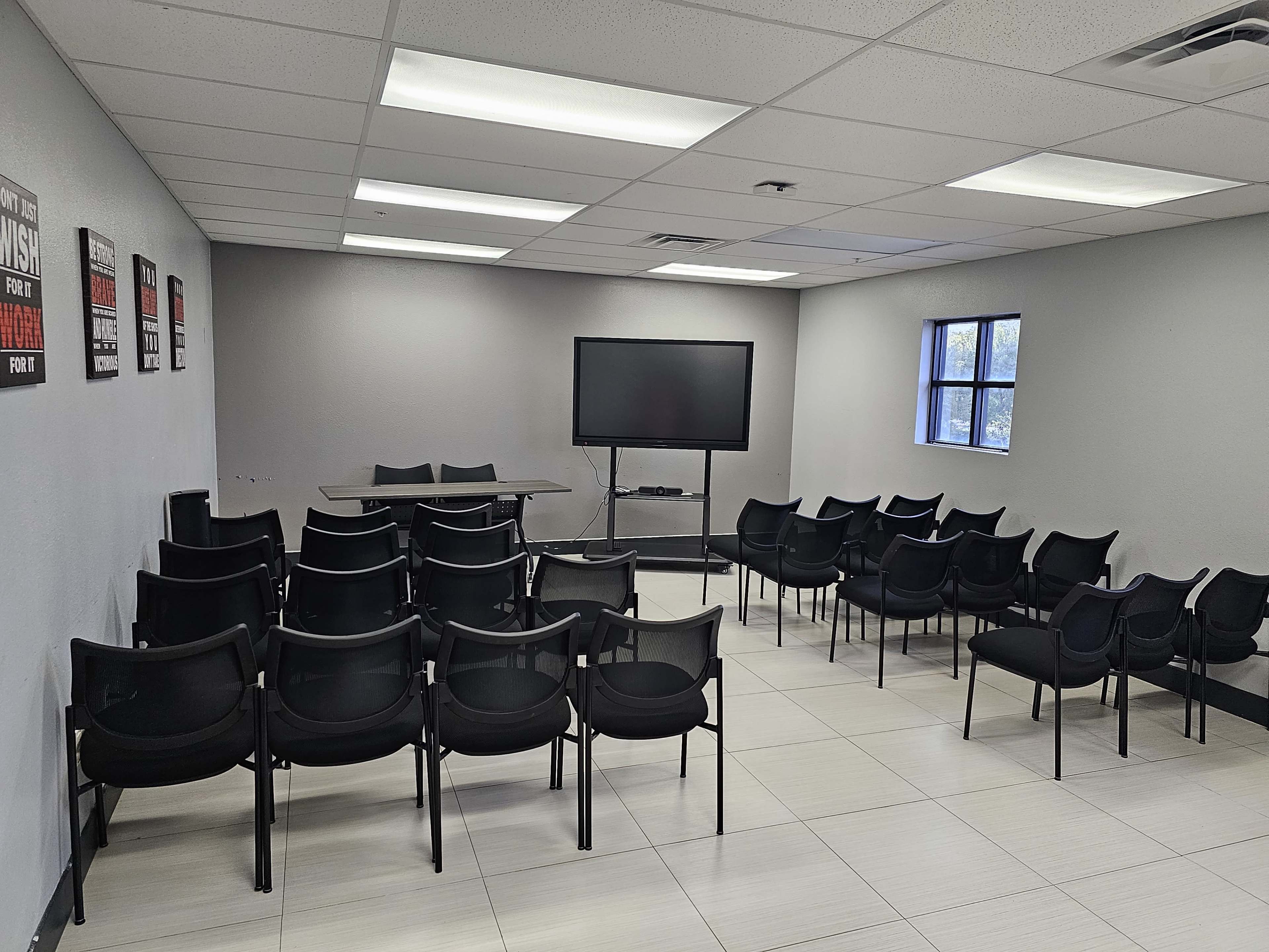 The image shows an empty meeting room with rows of black chairs facing a table and a wall-mounted screen.