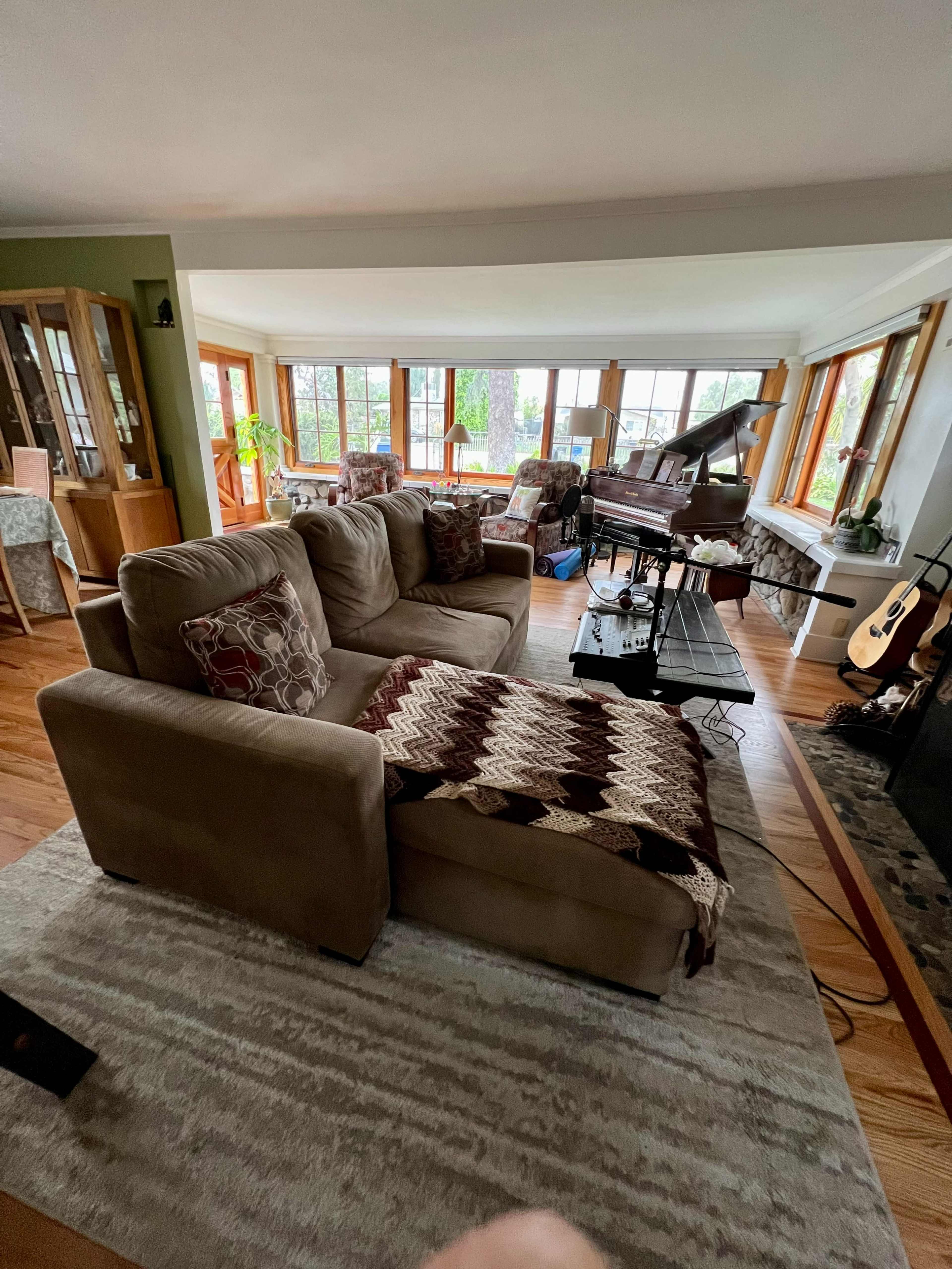The image shows a cozy living room with a beige sectional sofa, a grand piano, and large windows offering a view of greenery outside.